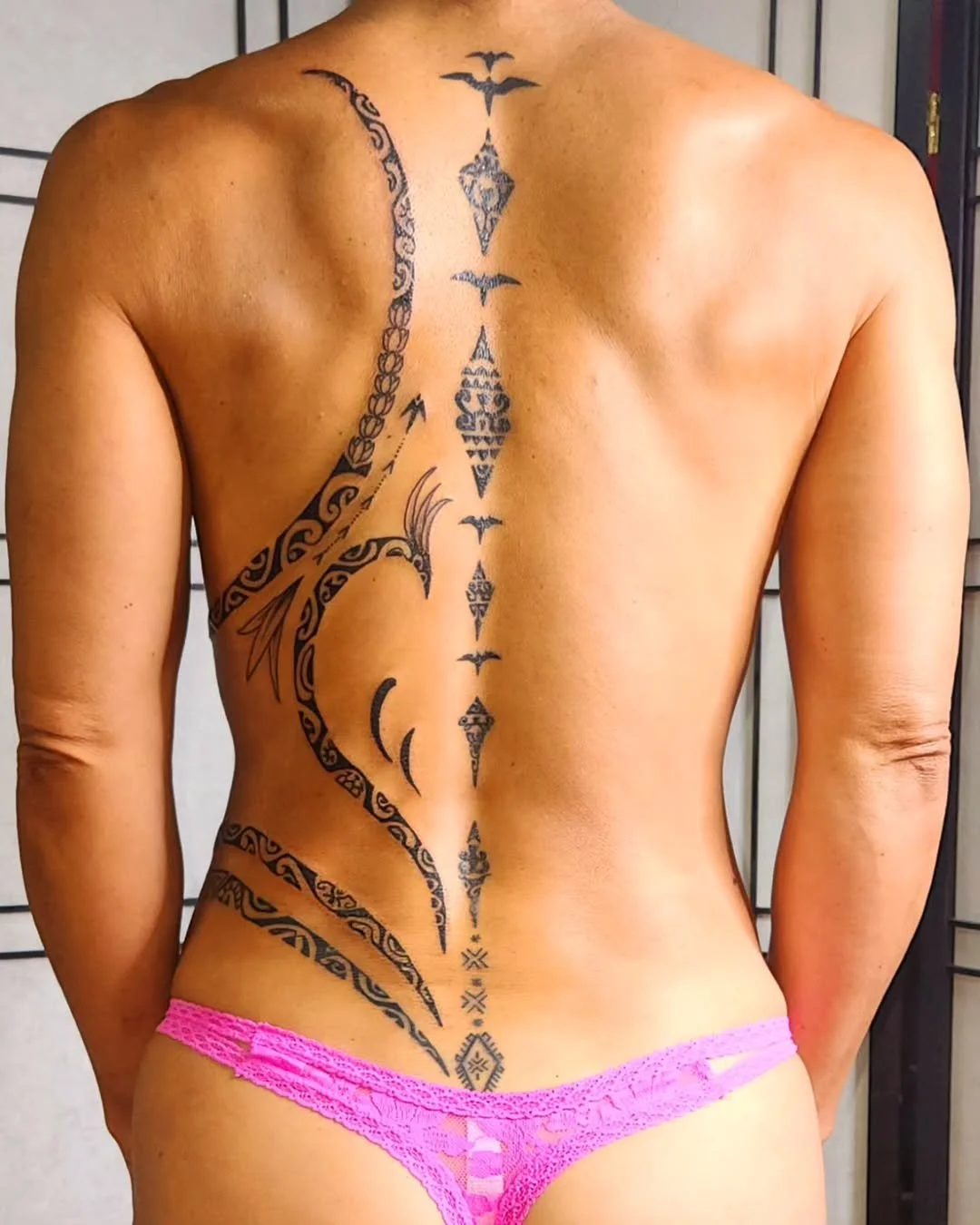 Back view of a person with a tribal-style tattoo running along the spine, with pink lace underwear.