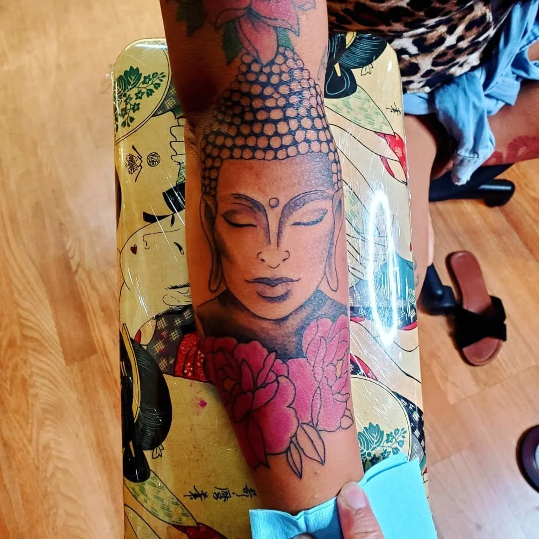 A tattoo of a serene Buddha face with closed eyes, detailed eyebrows, elongated earlobes, and floral elements on the arm.