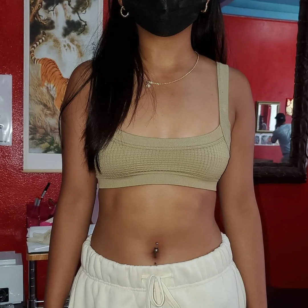 A woman with long dark hair wearing a black face mask, a beige cropped tank top, white sweatpants, a gold necklace, and earrings. She has a belly button piercing and is in a room with red walls, a tiger painting, and a mirror reflecting her back.