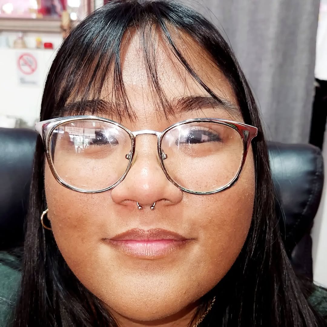 A woman with dark hair, wearing clear glasses, small hoop earrings, and a septum piercing, smiling while sitting in a chair in an indoor setting.