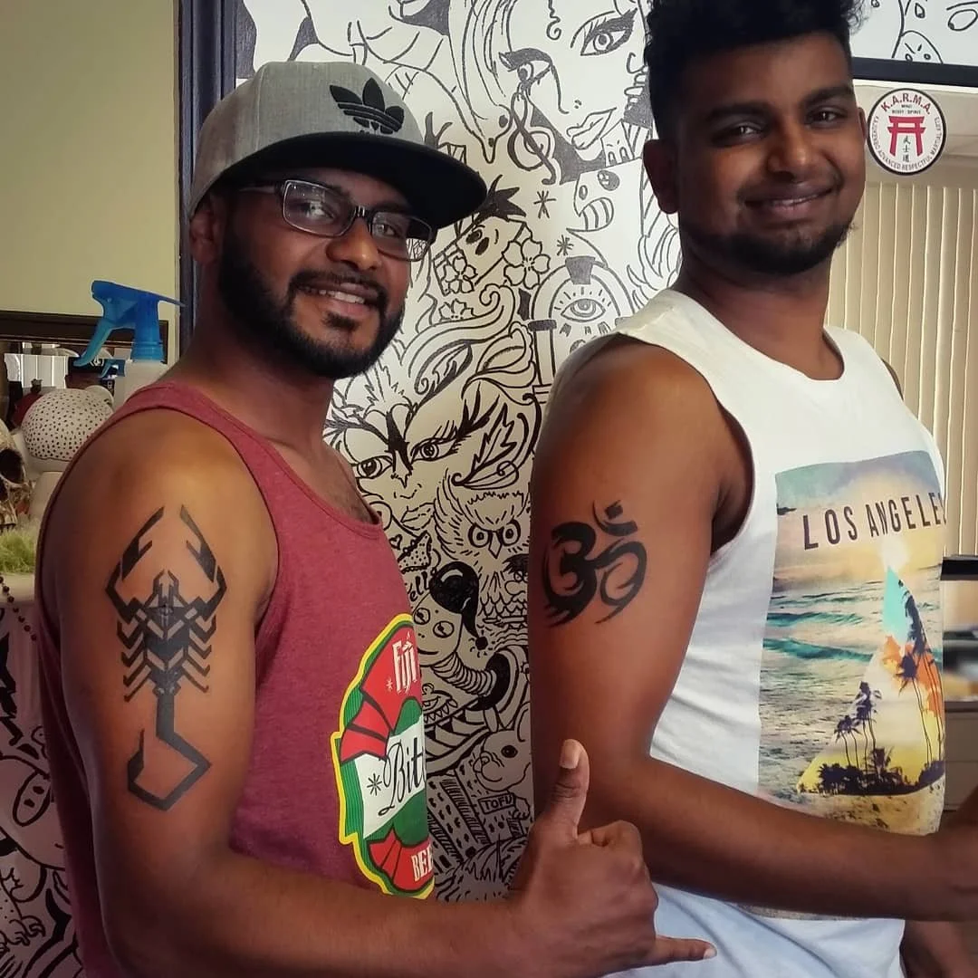 Two smiling men with tattoos on their arms, standing in front of a black and white mural with various artistic designs. One man is wearing a gray cap, glasses, and a red sleeveless shirt, giving a thumbs up. The other man is wearing a white sleeveles