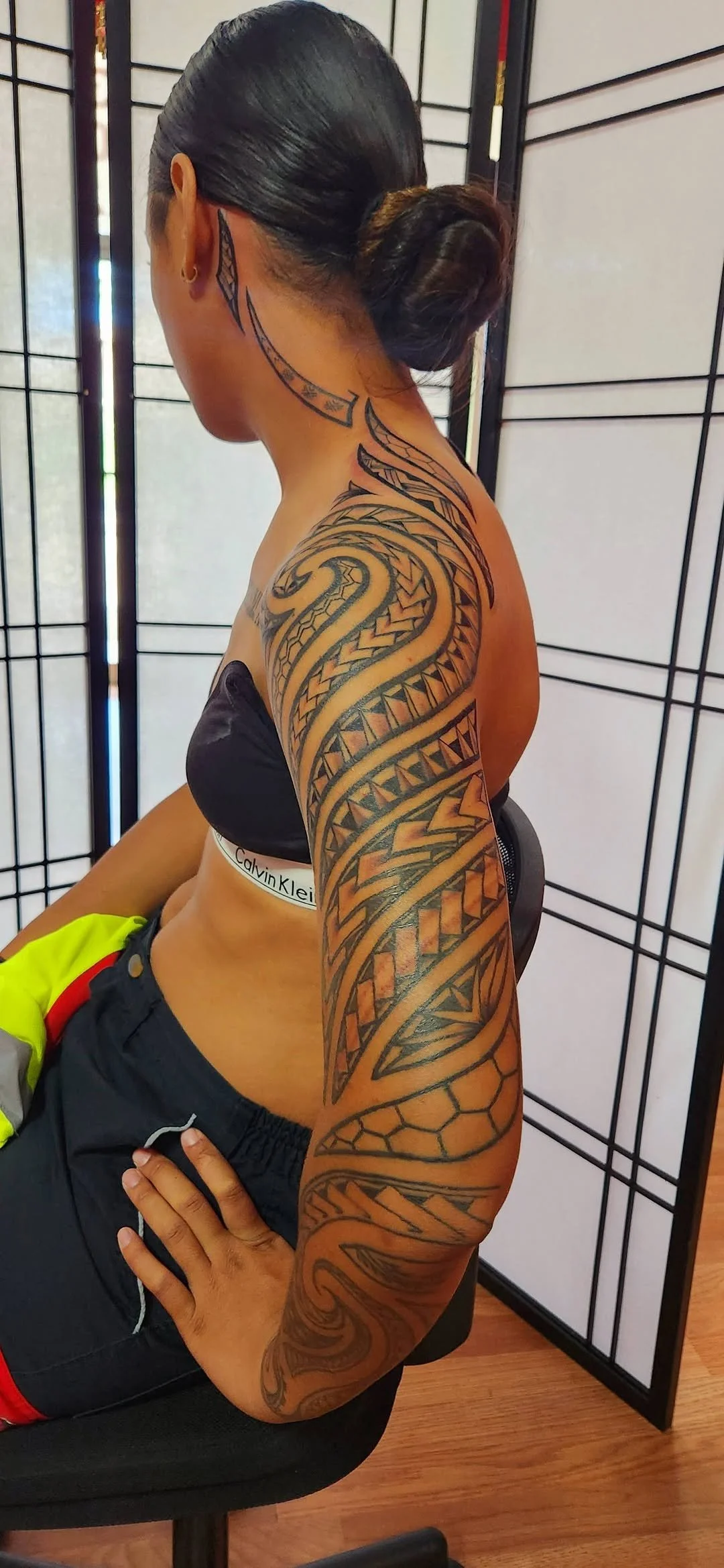 A woman with long black hair styled in a bun, wearing a black Calvin Klein sports bra and black pants with red accents, sitting on a chair in front of a folding room divider. She has extensive Polynesian-style black ink tattoos covering her neck, sho