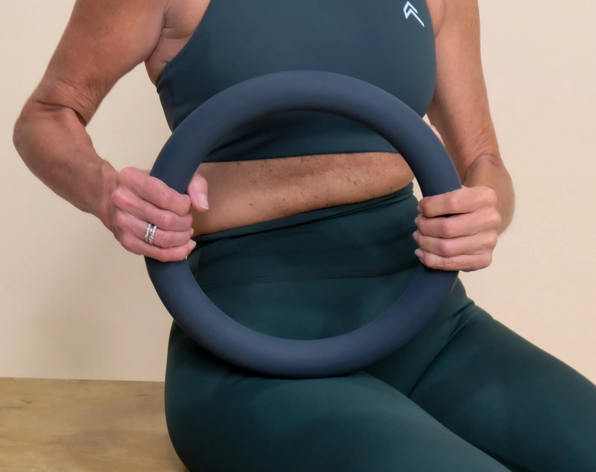 A woman sitting on a wooden surface, holding a gray exercise ring with both hands in front of her. She is wearing a dark green athletic sports bra and matching leggings.