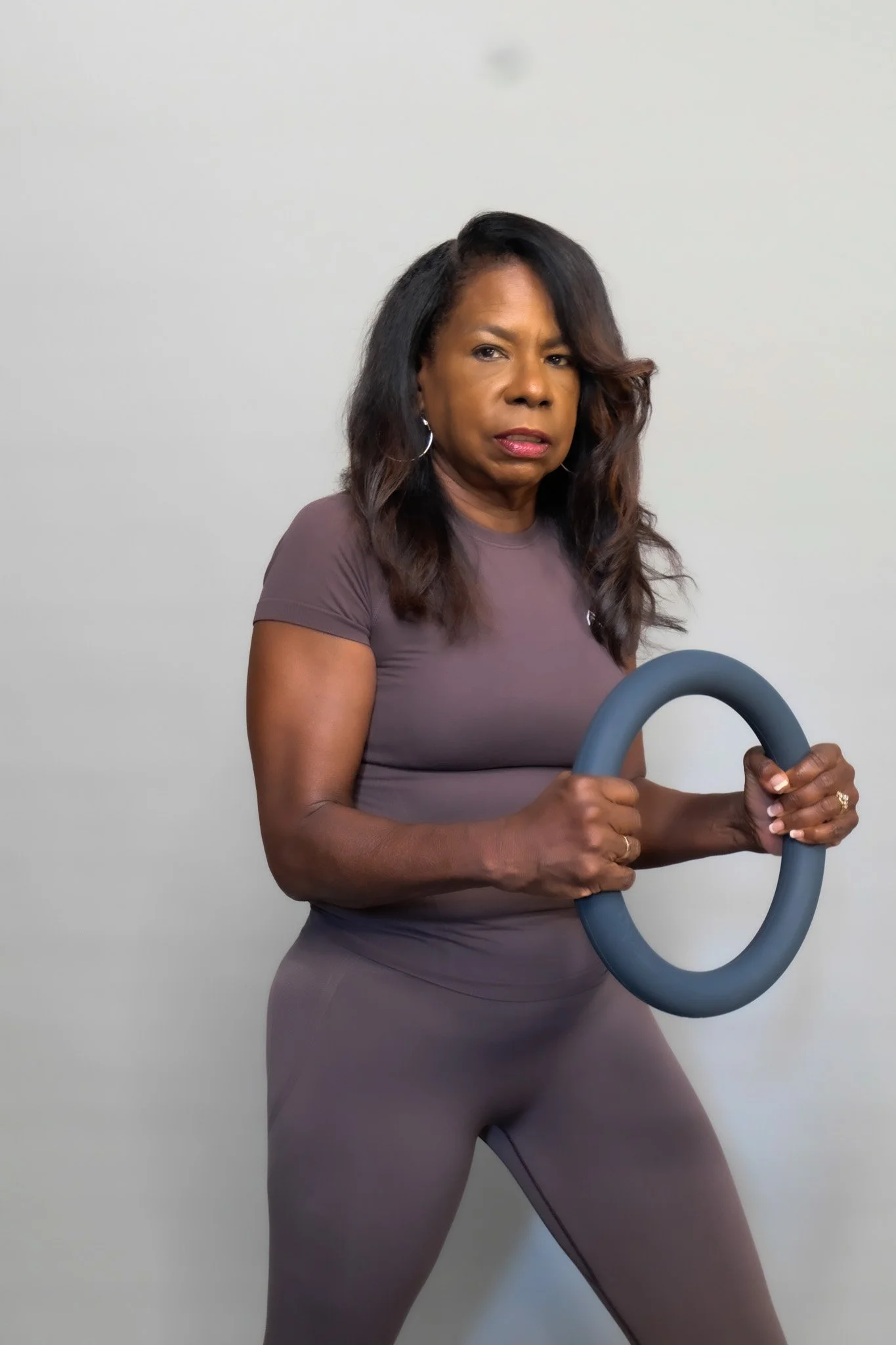 A woman dressed in athletic clothing holds a circular exercise ring with a serious expression, standing against a plain off-white wall.