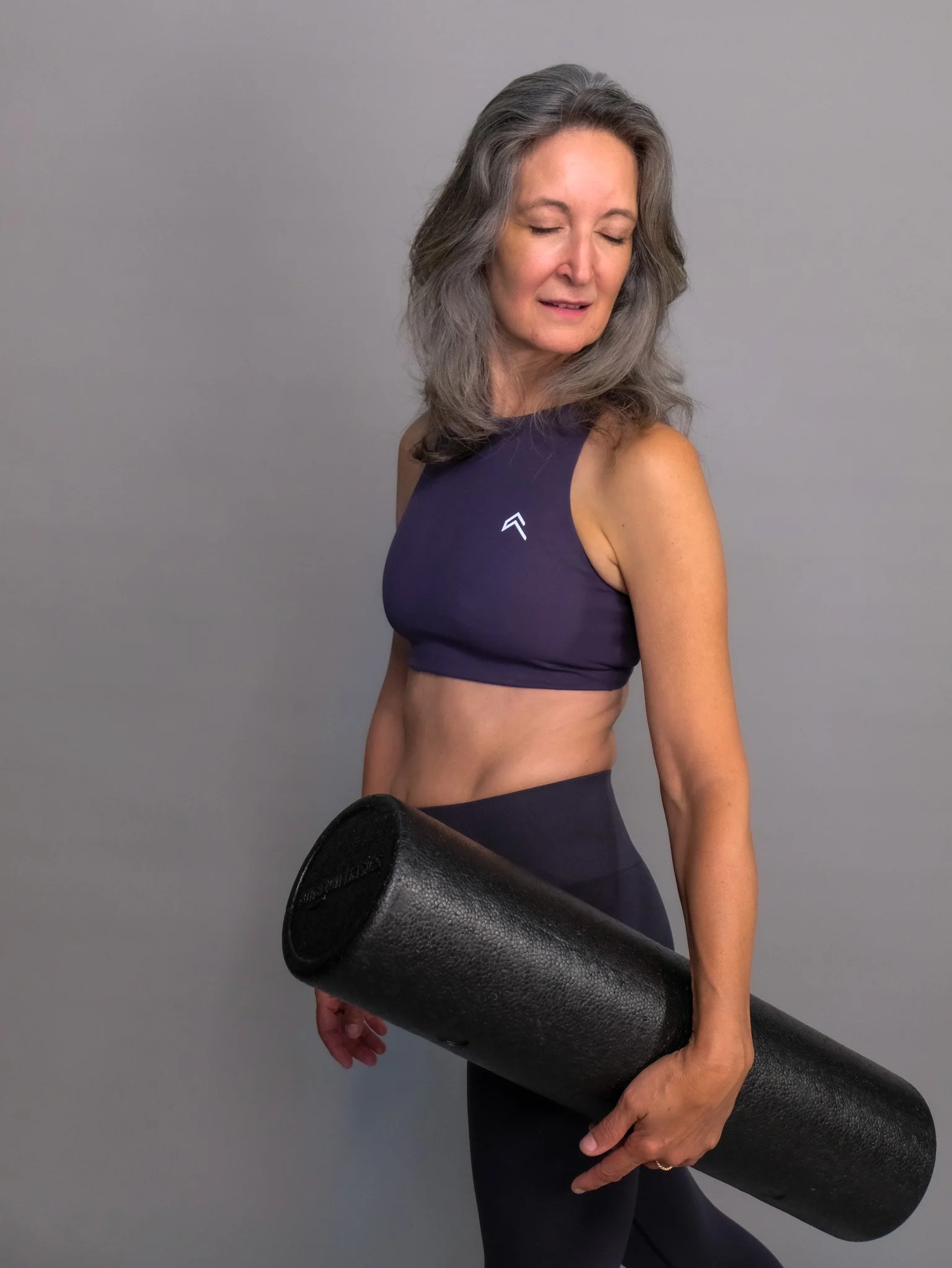 A woman with gray hair and wearing a purple sports top is holding a black foam roller.