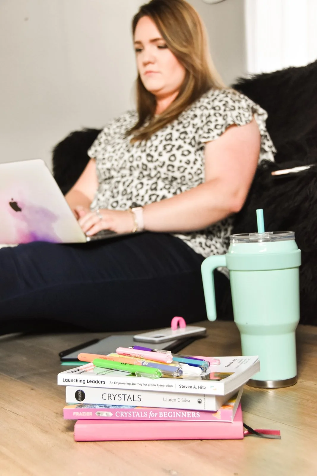 A woman sitting on the floor in front of a stack of books, using a laptop, with a reusable travel mug nearby.