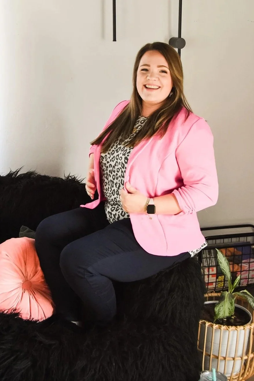 A woman with long brown hair, wearing a pink blazer and leopard print top, sitting on a black furry surface and smiling at the camera.