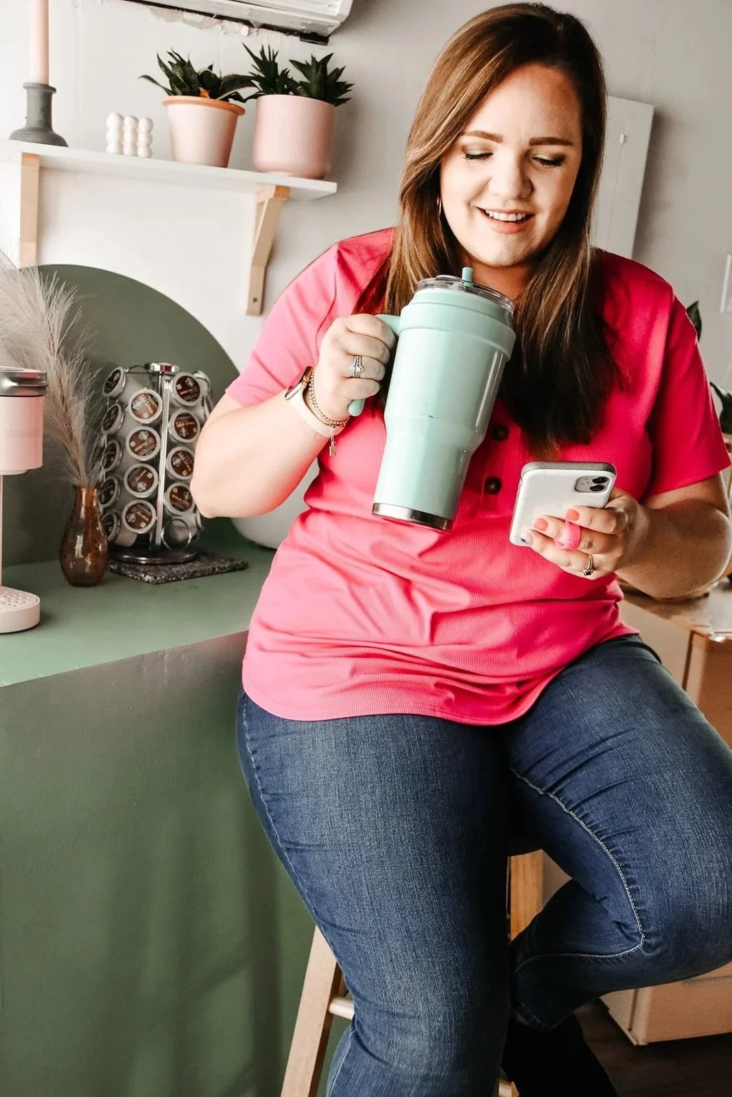 A woman sitting on a kitchen counter, holding a large mint-green tumbler and looking at her phone, wearing a pink shirt and blue jeans.