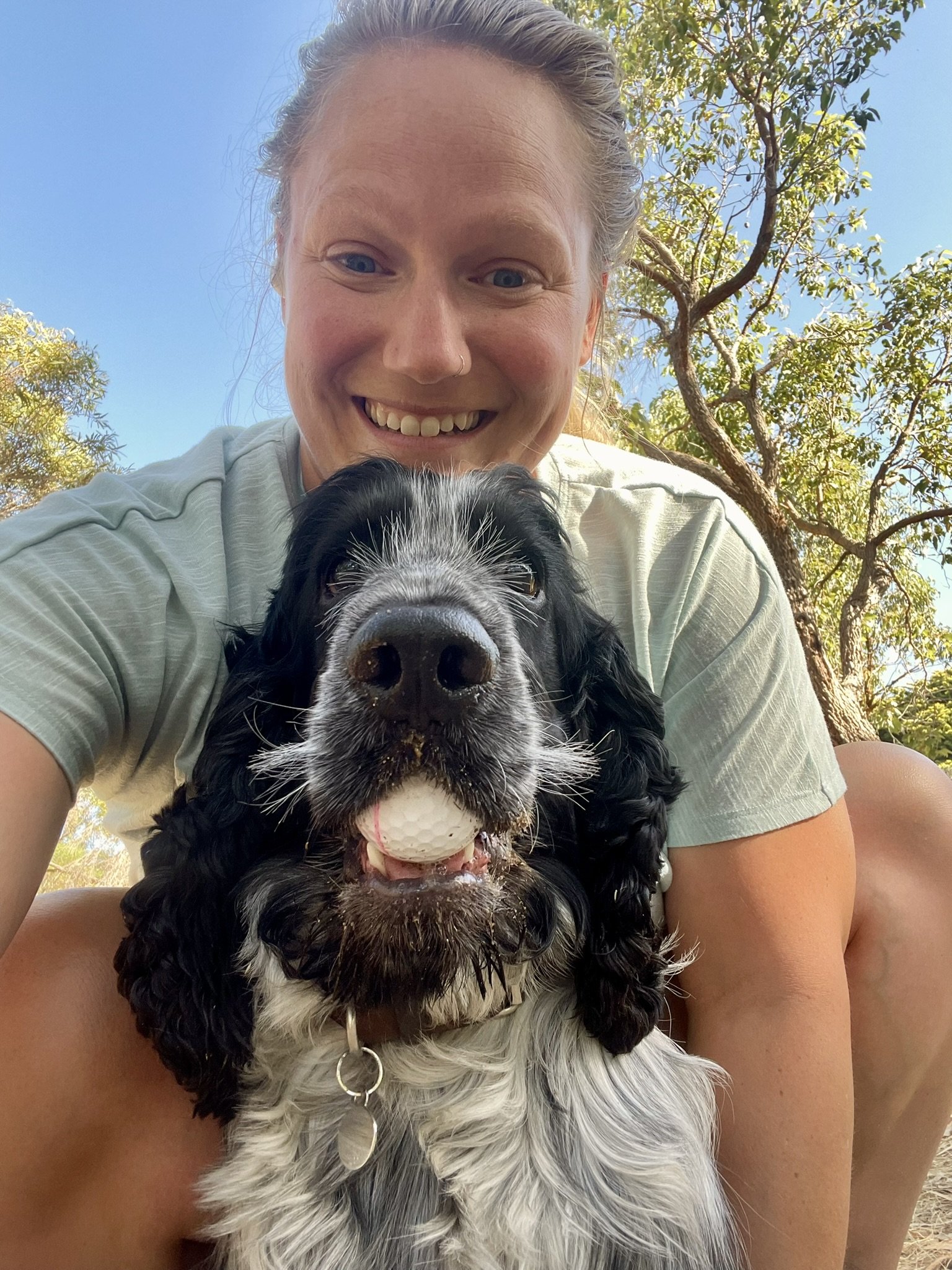 A woman taking a selfie with her black and white English Cocker Spaniel outdoors on a sunny day. The dog is holding a white golf ball in its mouth, and there are trees with green leaves behind them.