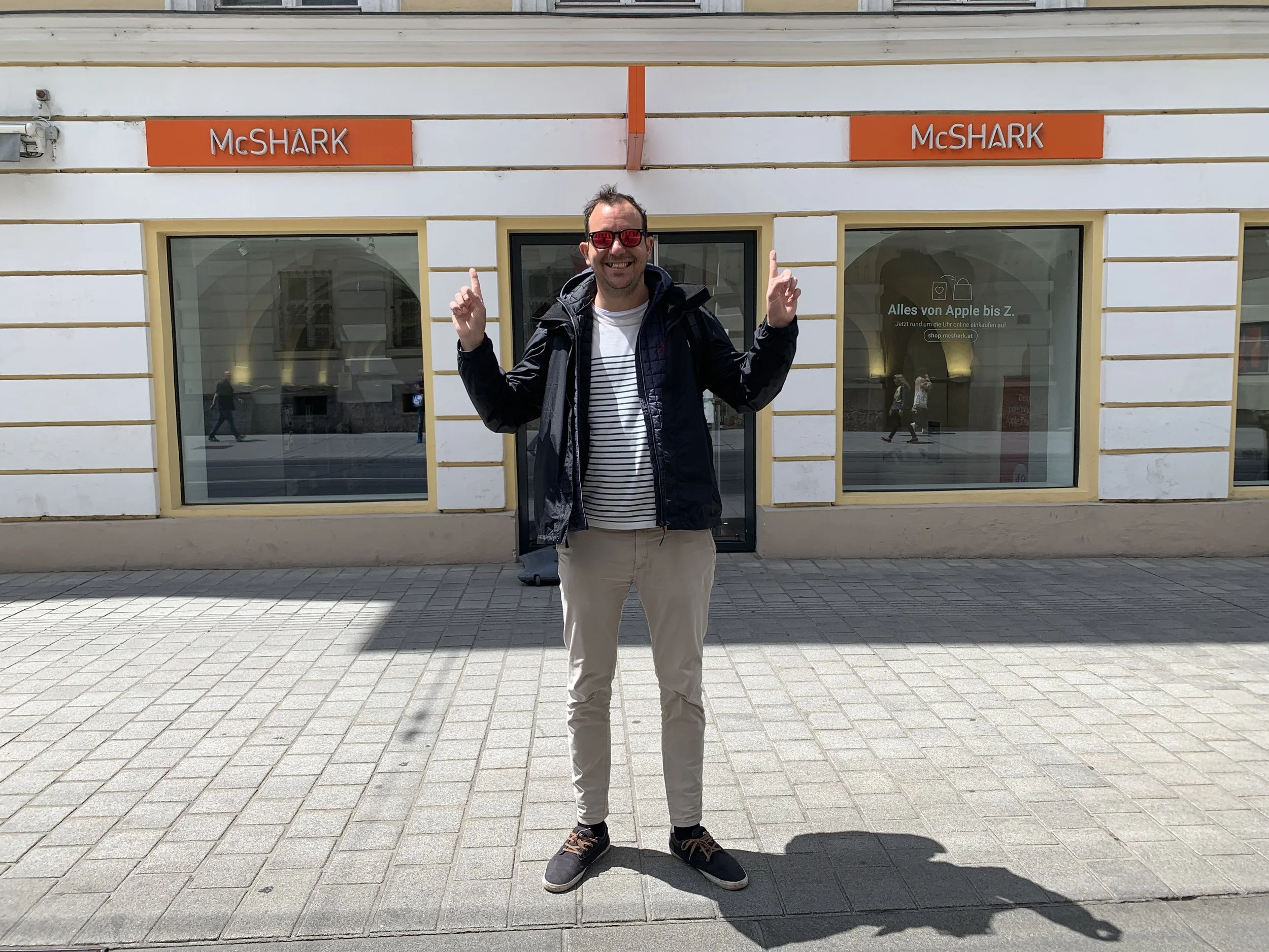 Hombre sonriendo, usando gafas de sol rojas, chaqueta negra, camisa blanca con rayas, pantalones beige y zapatos deportivos, posando con los brazos levantados frente a una tienda 'McShark' en una calle urbana