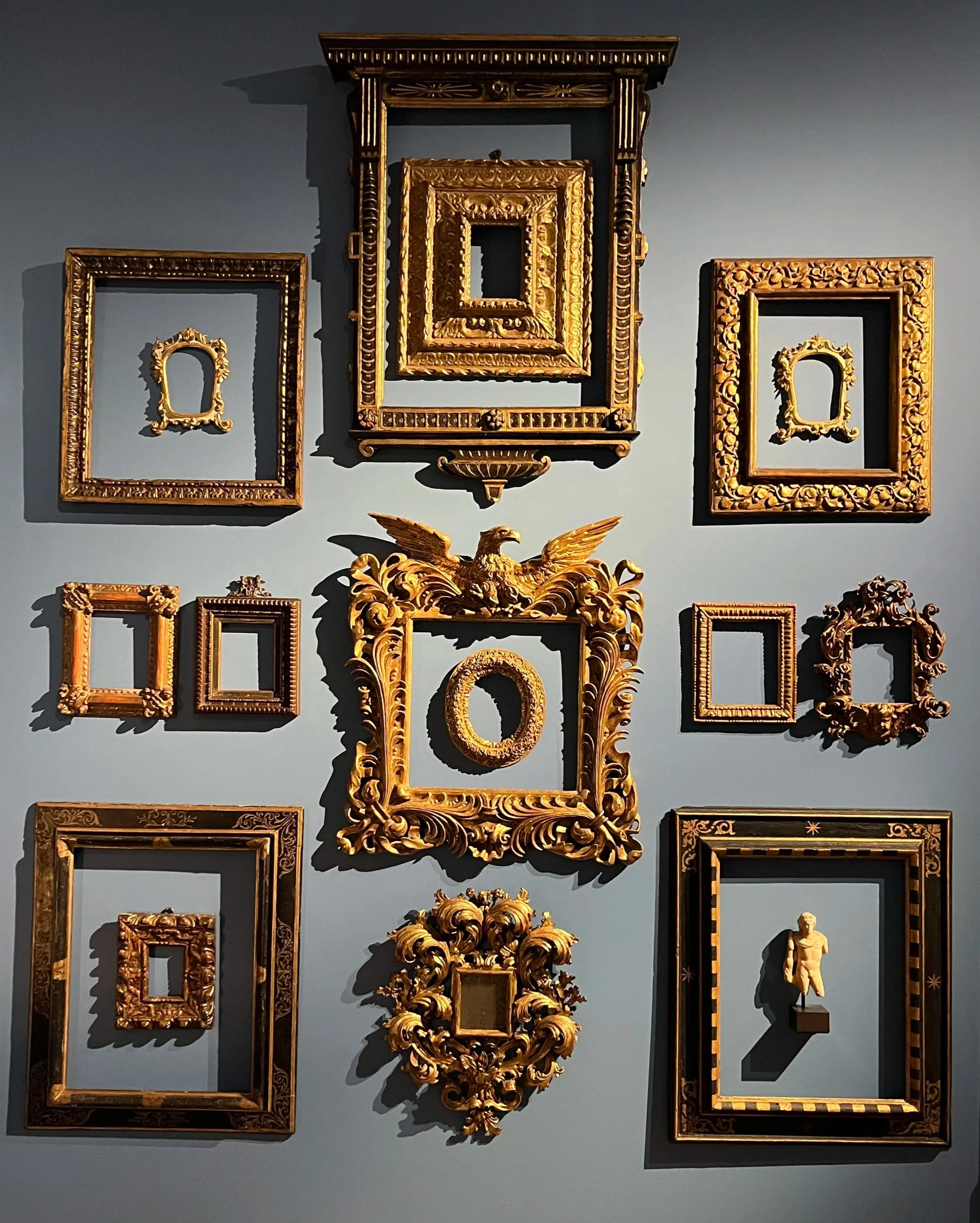 Conjunto de marcos dorados y un busto en una pared de color azul.