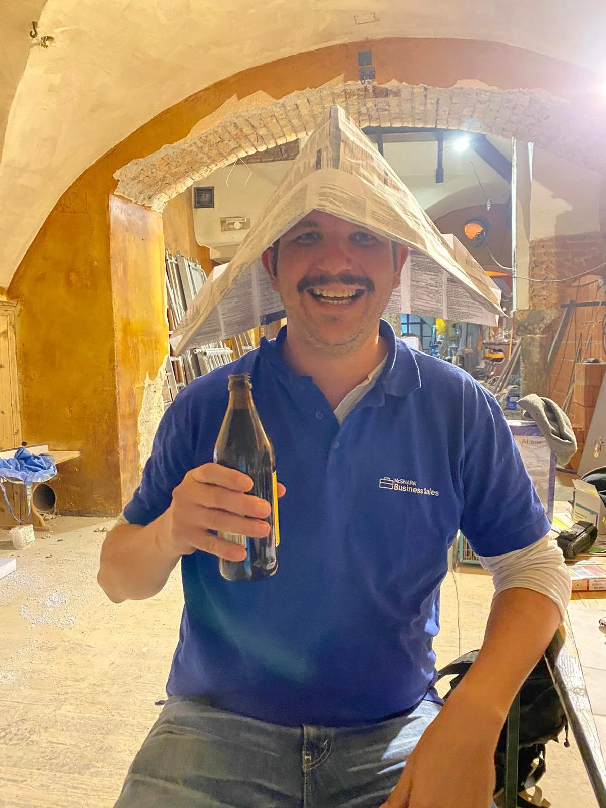 Hombre con sombrero de periódico, sonriendo y sosteniendo una botella en un espacio interior con arcos y paredes de ladrillo.