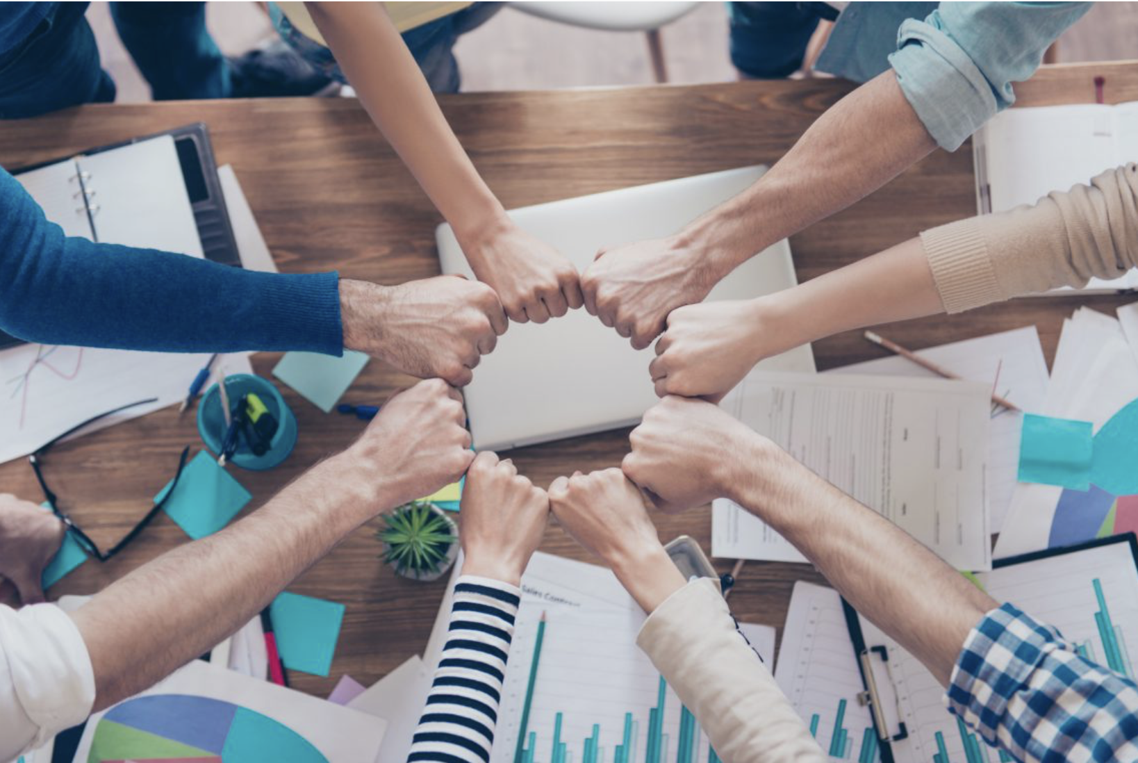 Varias personas uniendo sus manos en un gesto de colaboración sobre una mesa de trabajo con documentos, gráficas y objetos de oficina.