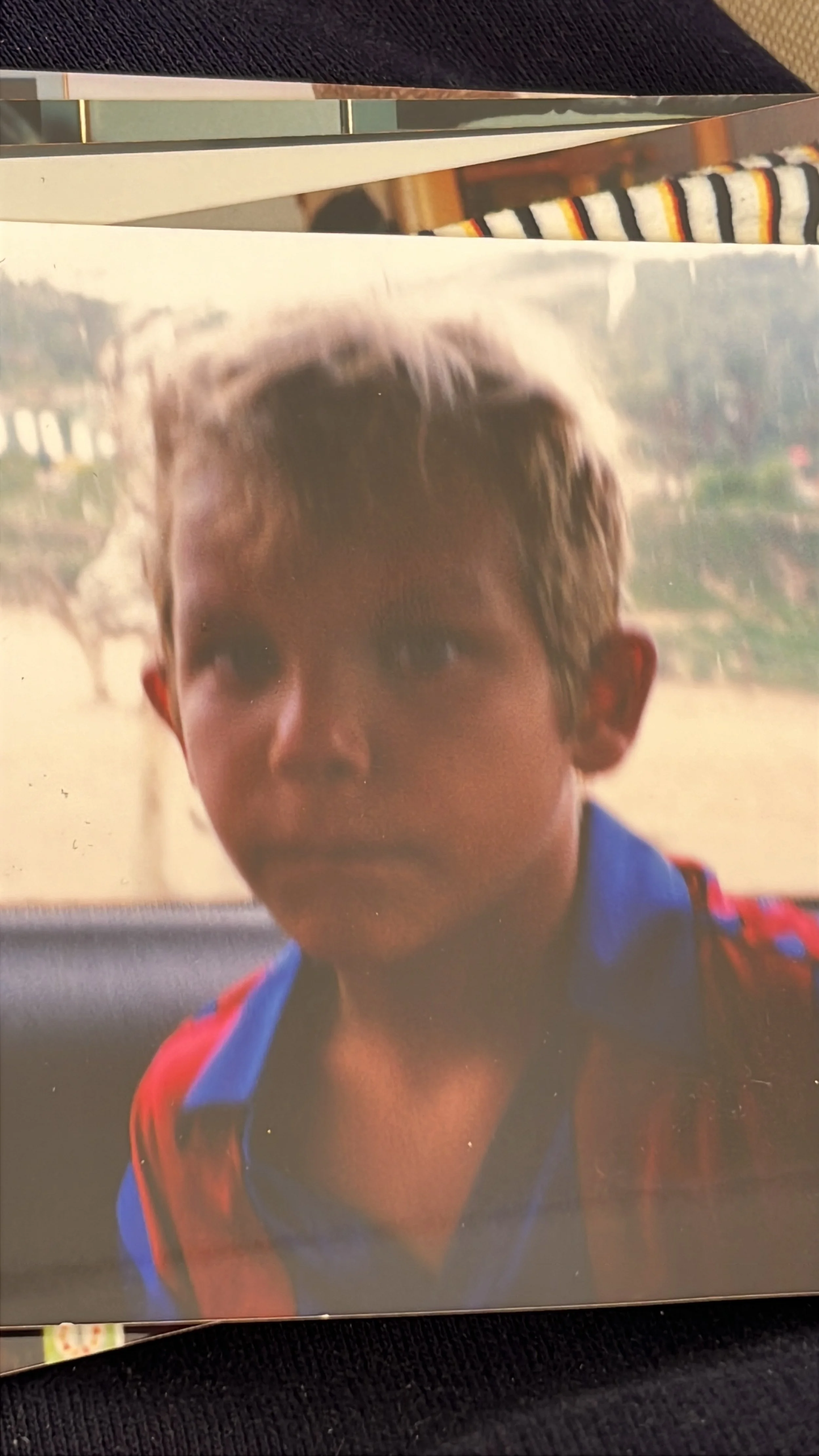 Niño con cabello rubio y ojos claros, usando camiseta azul y chaqueta roja con negro oscuro, en el interior de un vehículo.