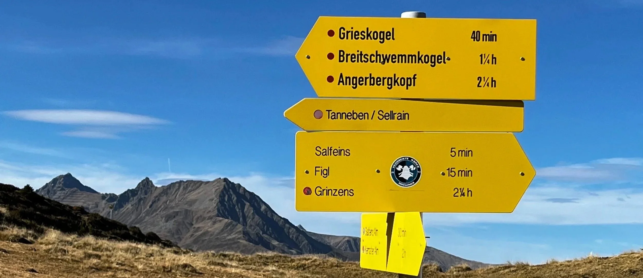 Señalización de senderismo con destinos y tiempos, en un paisaje montañoso con cielo azul y nubes.