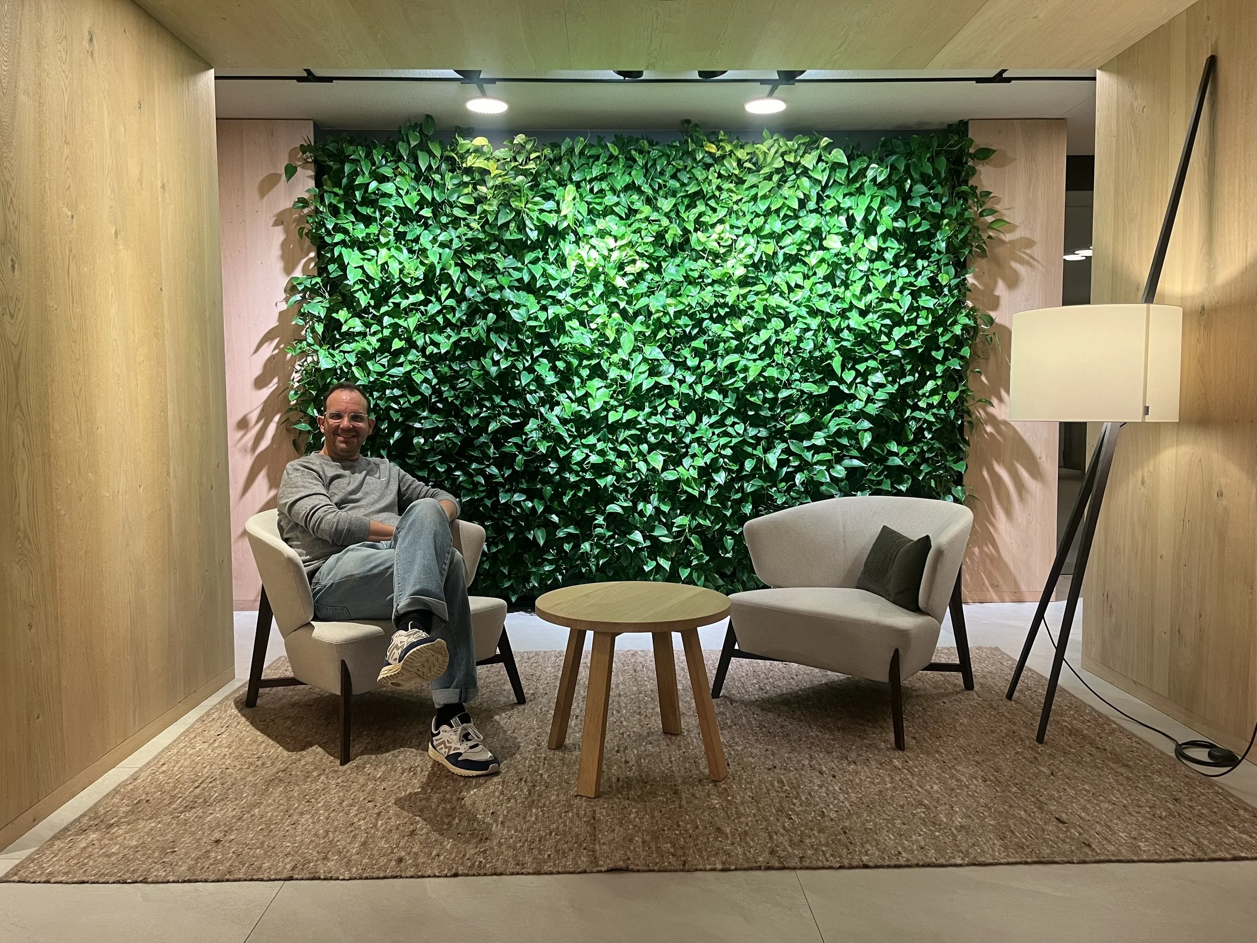 Hombre sentado en sala moderna con paredes de madera, alfombra, sofá de diseño y pared decorativa de plantas verdes.