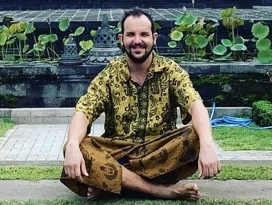 Hombre sentado en posición de meditación o yoga junto a un estanque con plantas acuáticas, vestido con ropa tradicional con patrones dorados y marrones.