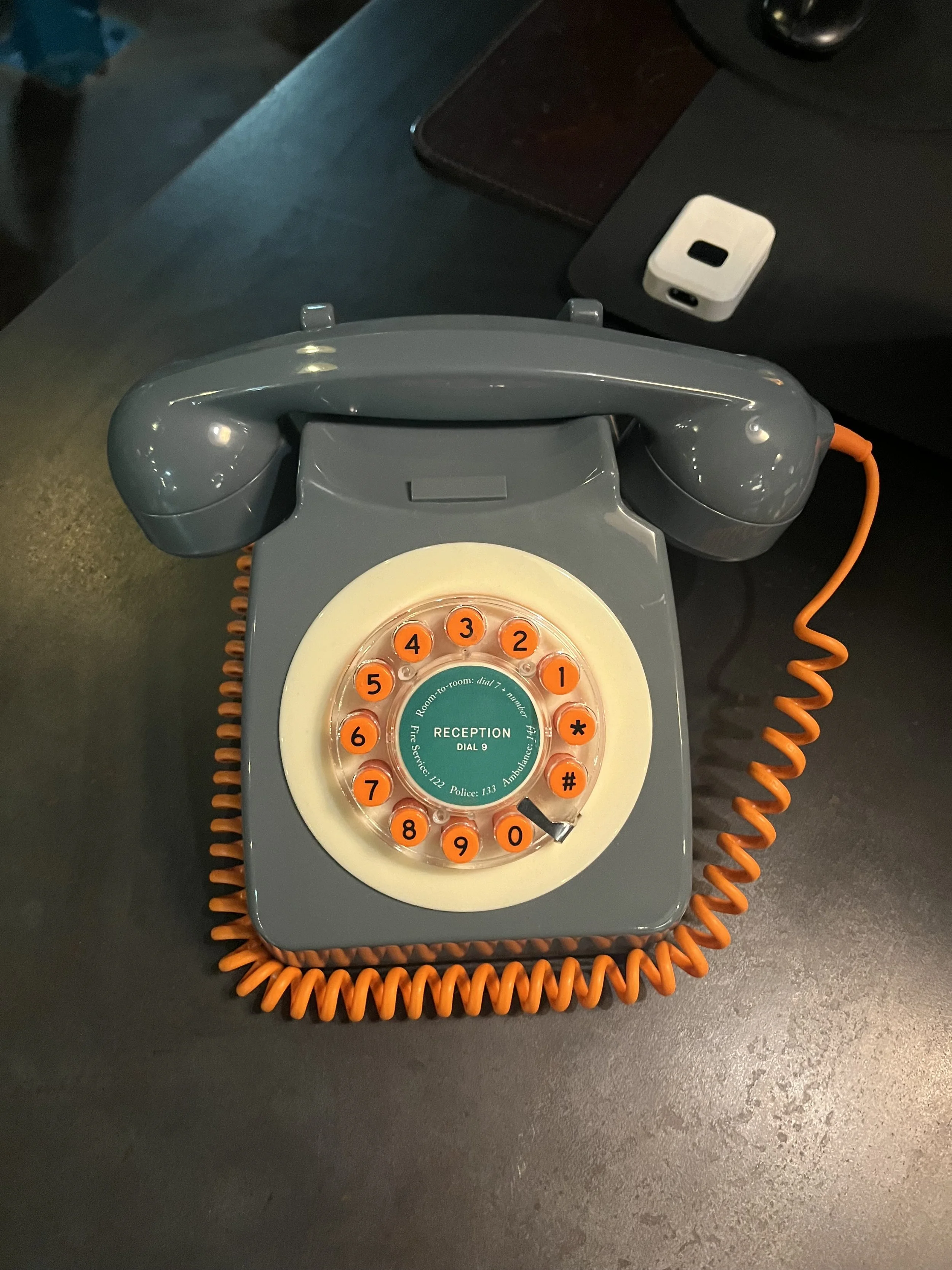 Teléfono de disco clásico de color gris con cables naranjas, colocado sobre una mesa negra junto a otros objetos.