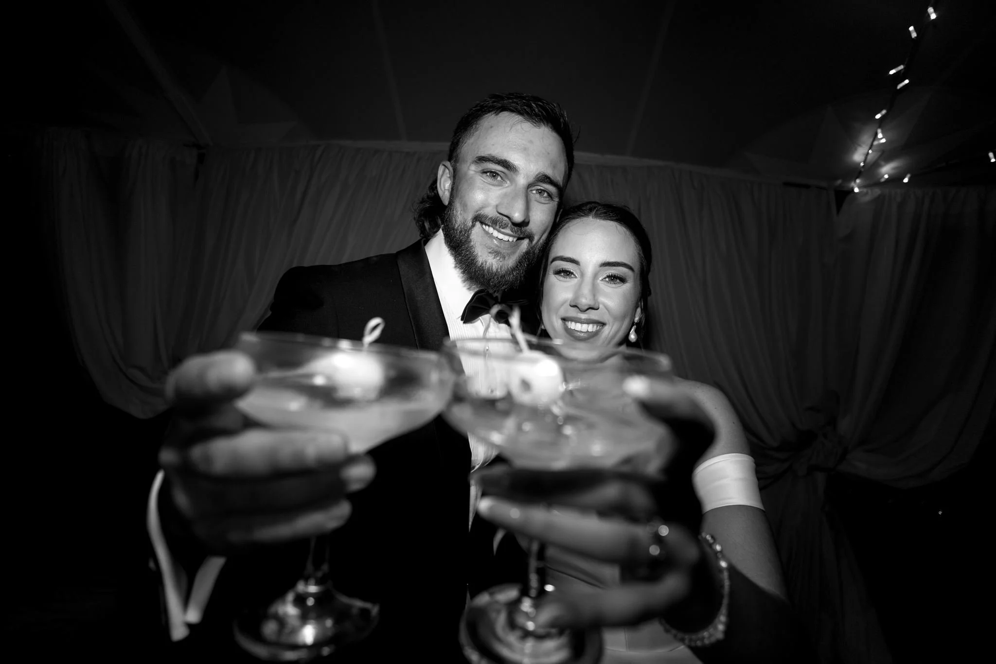 A happy couple in formal attire raising glasses for a toast at a celebration, with the focus on their smiling faces and drinks, in black and white.