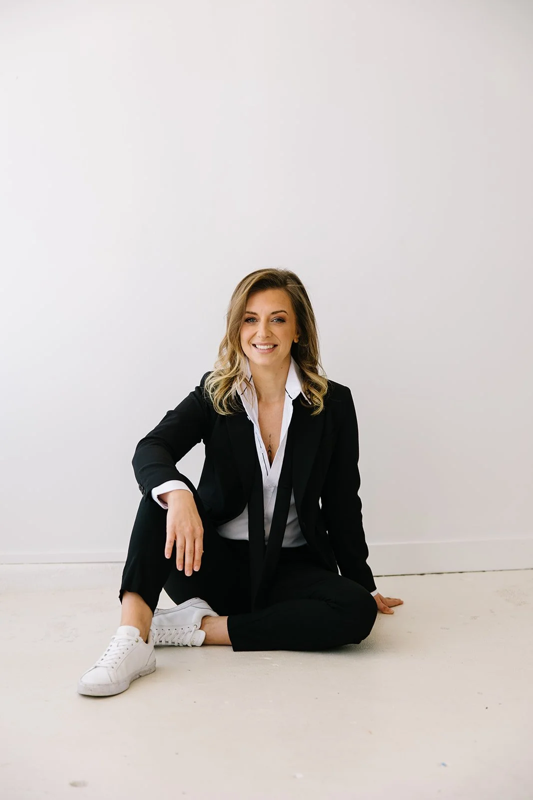 A woman with blonde hair sitting on the floor against a plain white wall, dressed in a black suit and white sneakers, smiling at the camera.