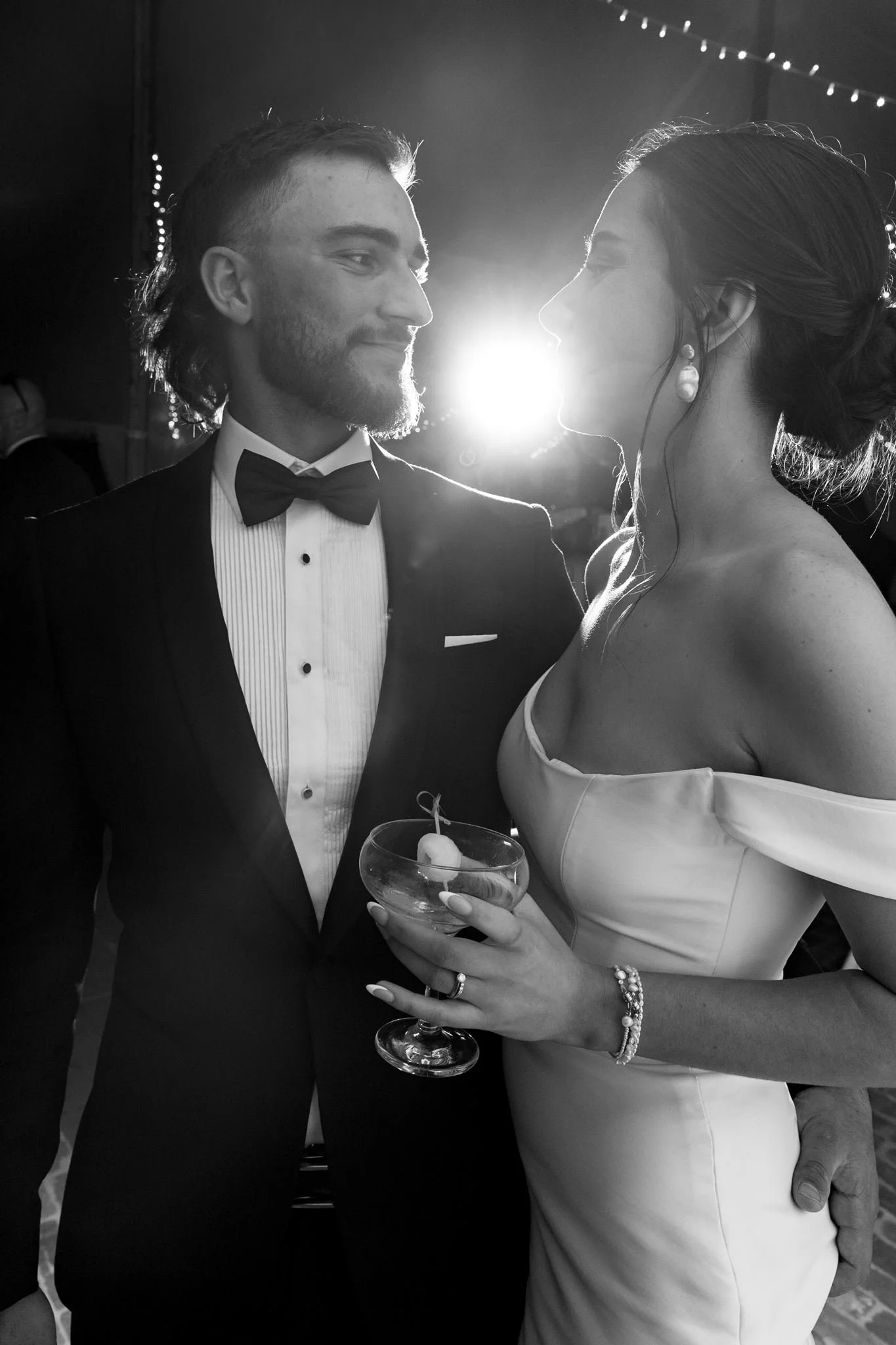 A black-and-white photo of a couple at a formal event, staring into each other's eyes, standing close. The man wears a tuxedo with a bow tie and the woman is in a strapless dress holding a cocktail glass. The woman has jewelry, including earrings and a bracelet. Bright backlighting creates a glow around them.