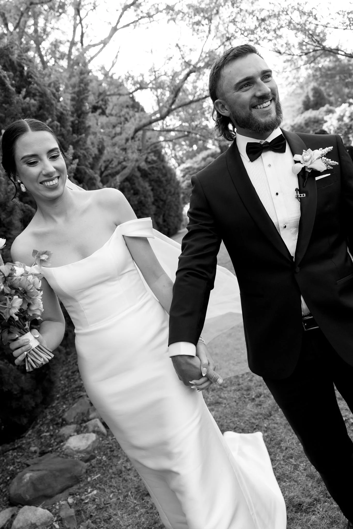 A black and white photo of a smiling bride and groom holding hands outdoors, with trees in the background.
