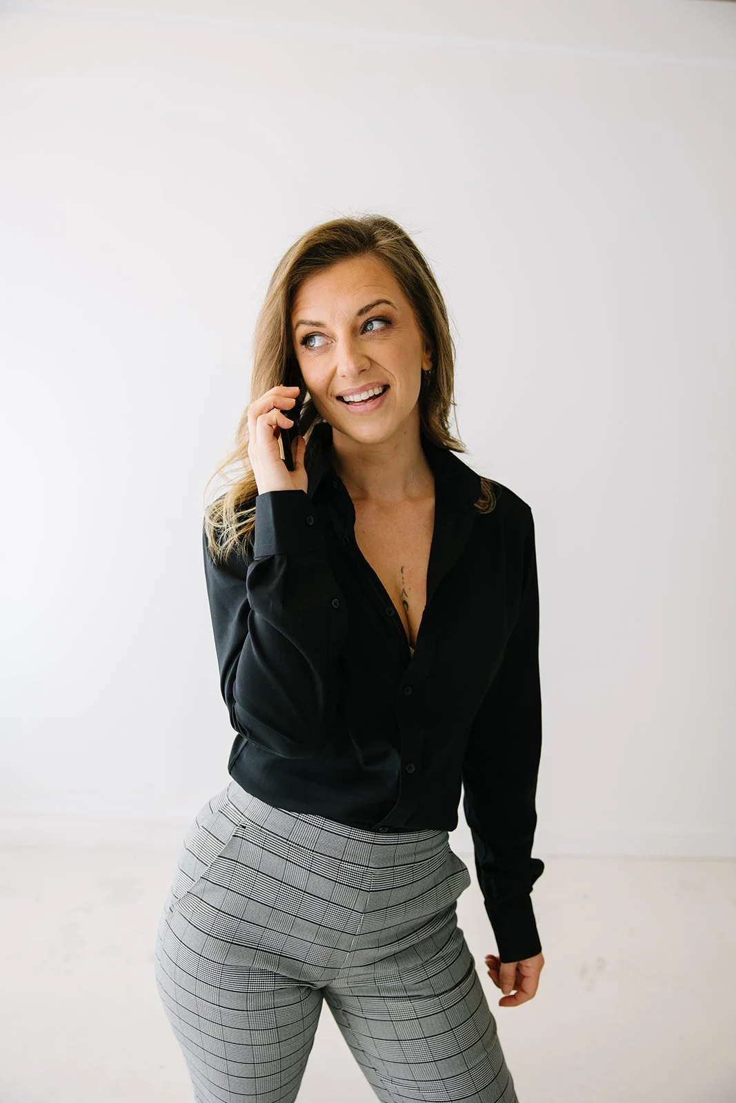 A woman with light brown hair, wearing a black button-up shirt and gray plaid pants, talking on her cellphone with a slight smile, standing against a plain white background.