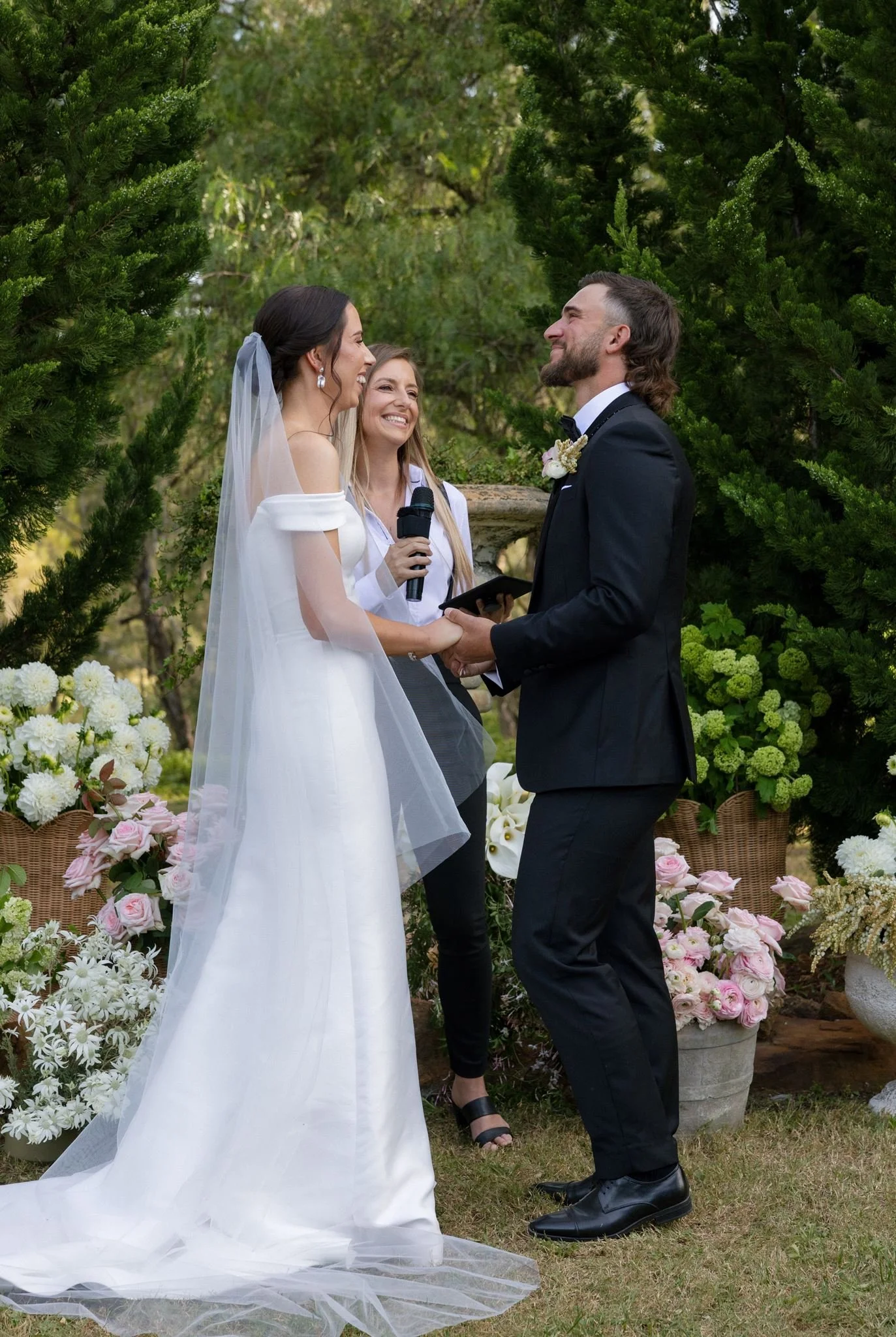 A wedding ceremony outdoors with a bride and groom holding hands and smiling at each other, surrounded by greenery and floral arrangements.