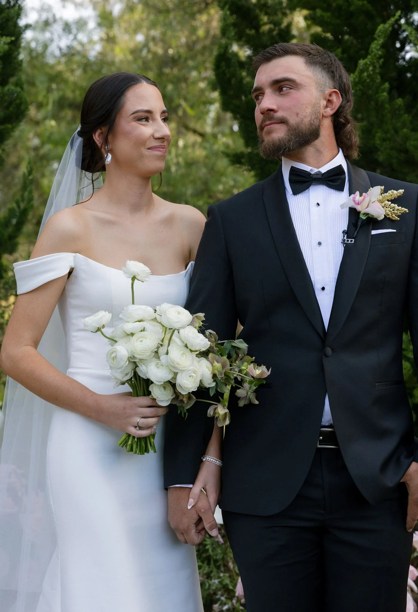 A bride holding a bouquet of white flowers standing next to a groom in a black tuxedo with a bow tie, both outdoors with green trees in the background.