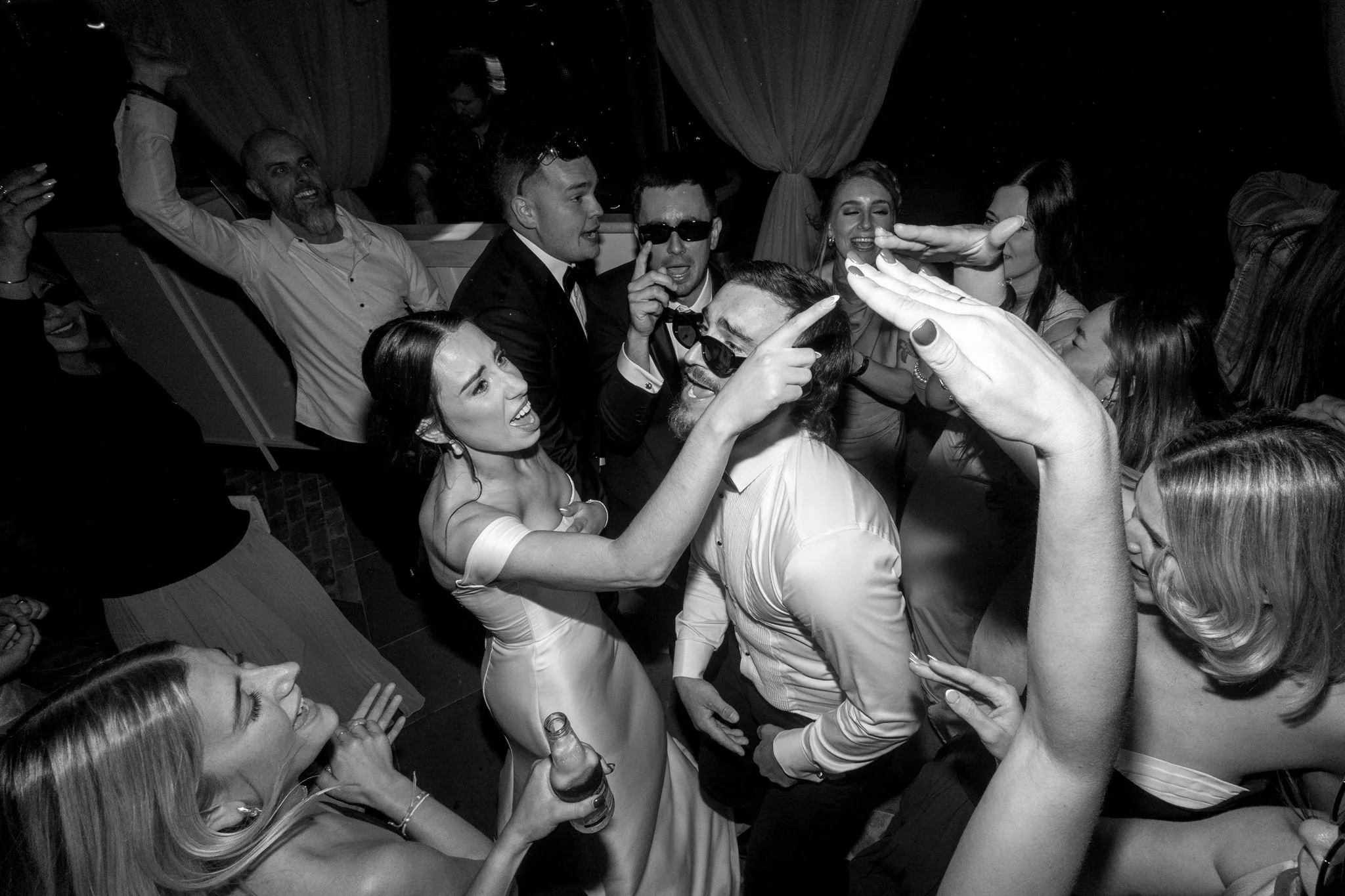 Group of people dancing and celebrating at a lively party or celebration, with some individuals making gestures and smiling.