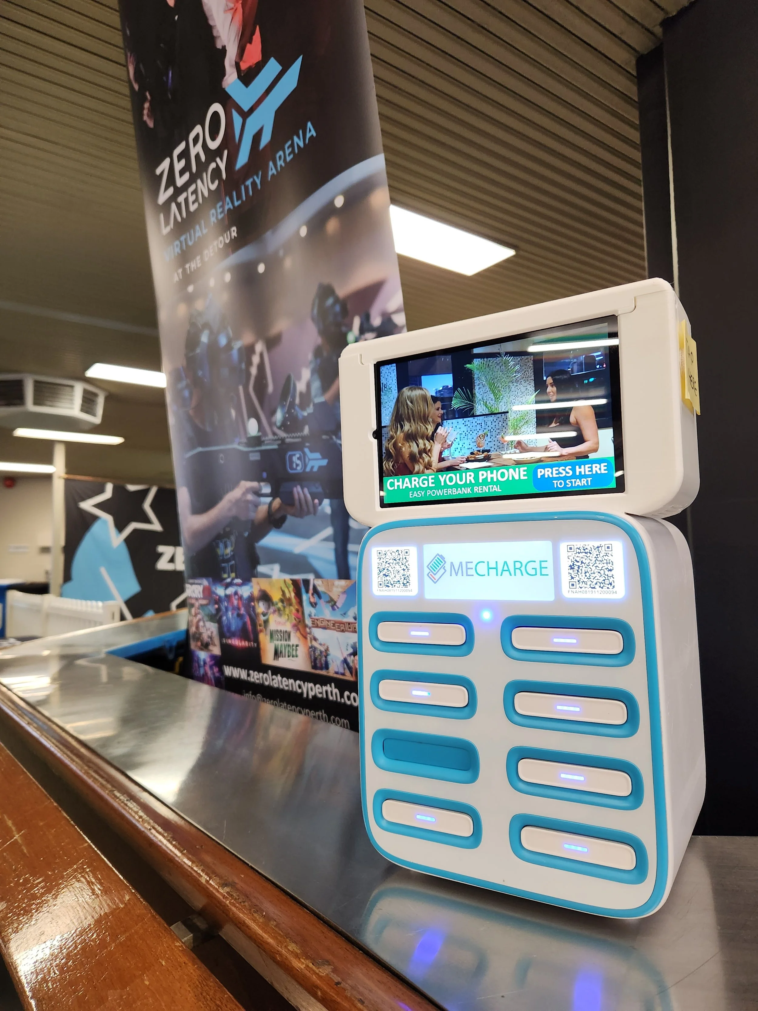 A phone charging station with a small screen displaying an ad, inside a gaming or entertainment center with a banner for Zero Latency Virtual Reality Arena in the background.
