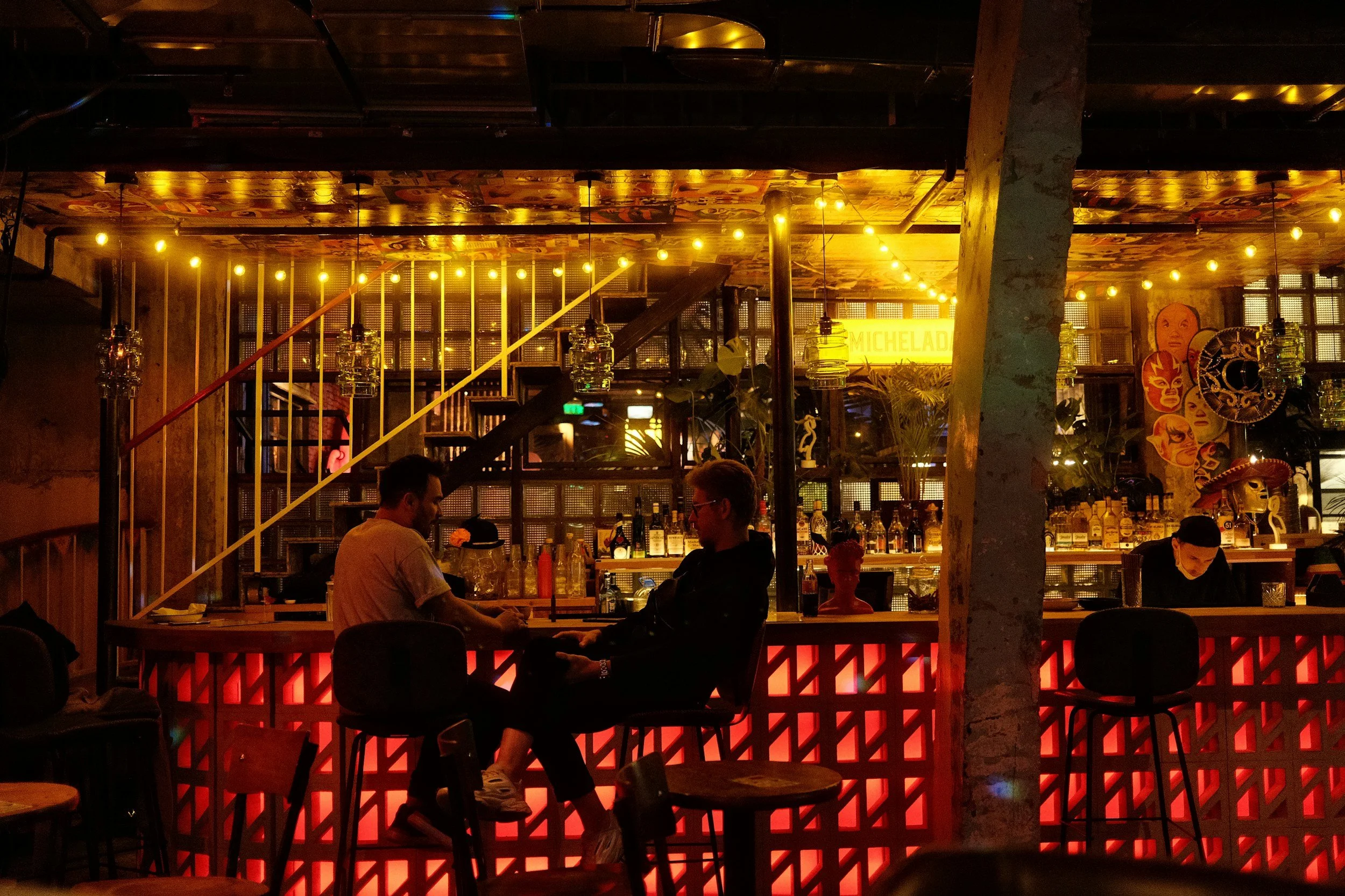 A dimly lit bar with two men sitting and talking at the counter, illuminated by warm yellow and orange string lights, with shelves of bottles and decorative masks on the wall in the background.