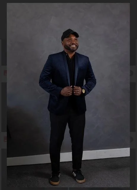 A man smiling confidently, wearing a dark blue blazer, black shirt, black pants, sneakers, a black cap, and a wristwatch, standing against a gray textured wall.