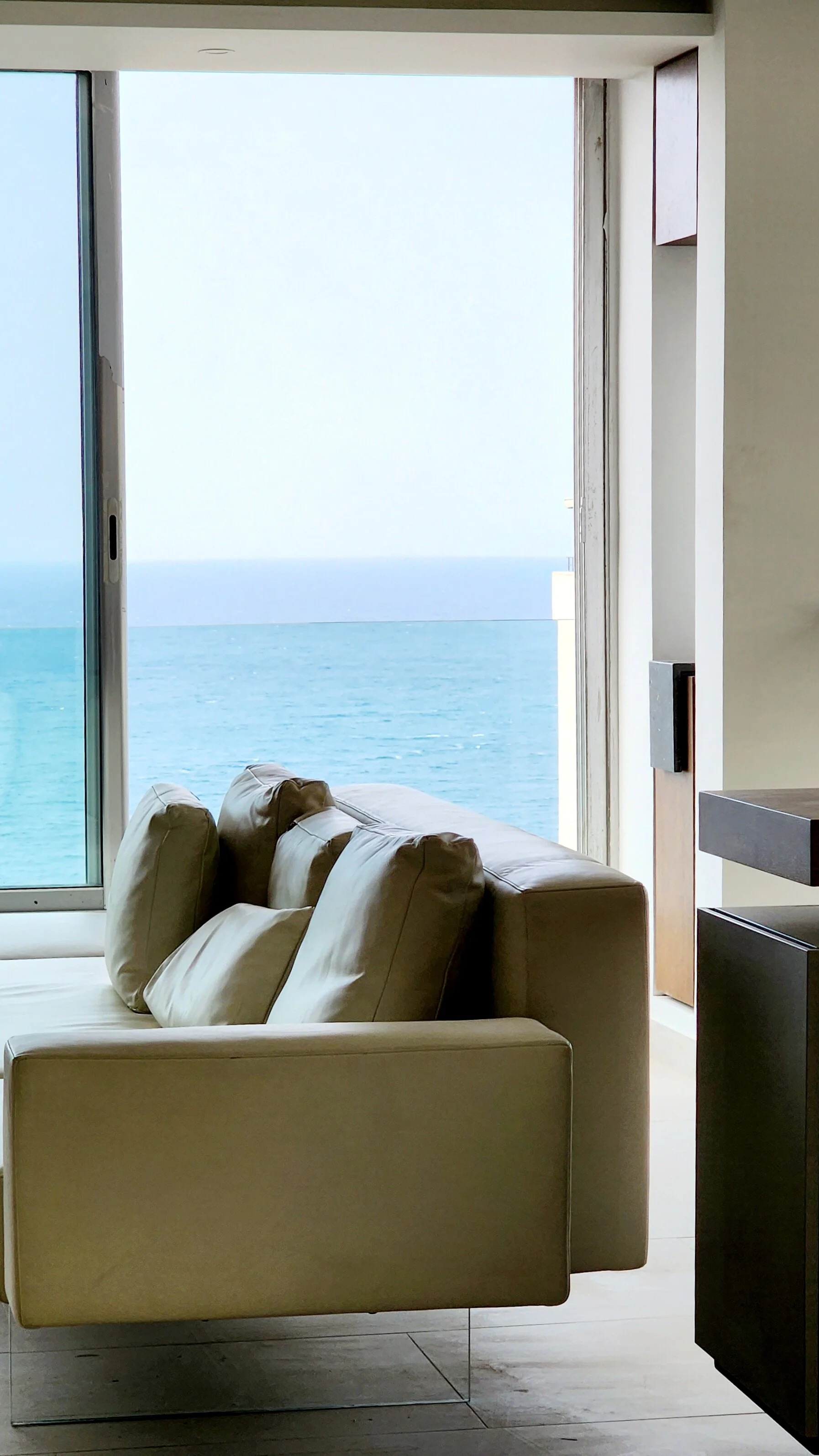 Living room with beige sofa facing large glass sliding door open to ocean view, with blue water and sky visible.