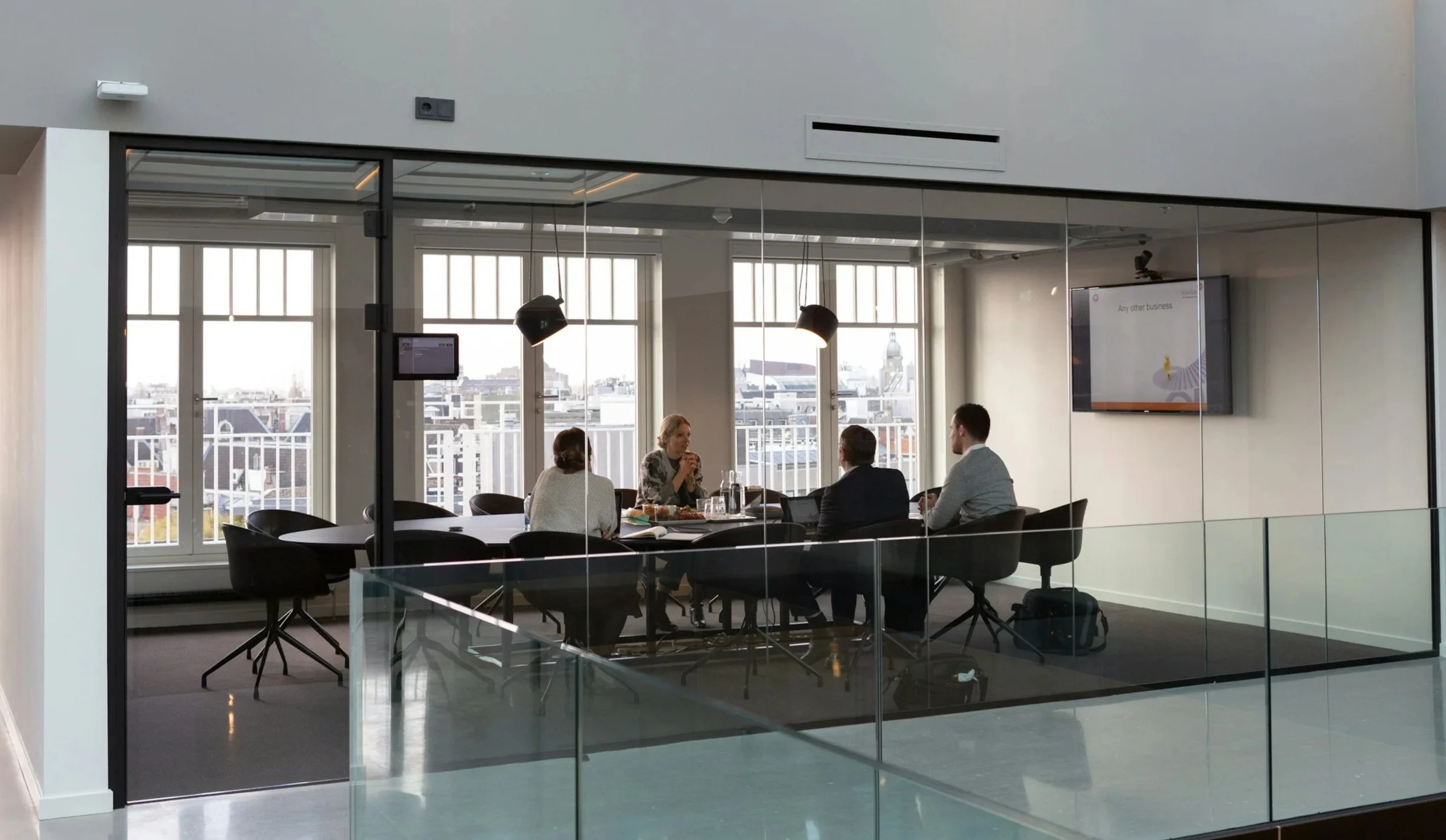 A modern conference room with four people seated around a table, engaging in a discussion. The room has large windows with city views, black office chairs, and a wall-mounted video screen.
