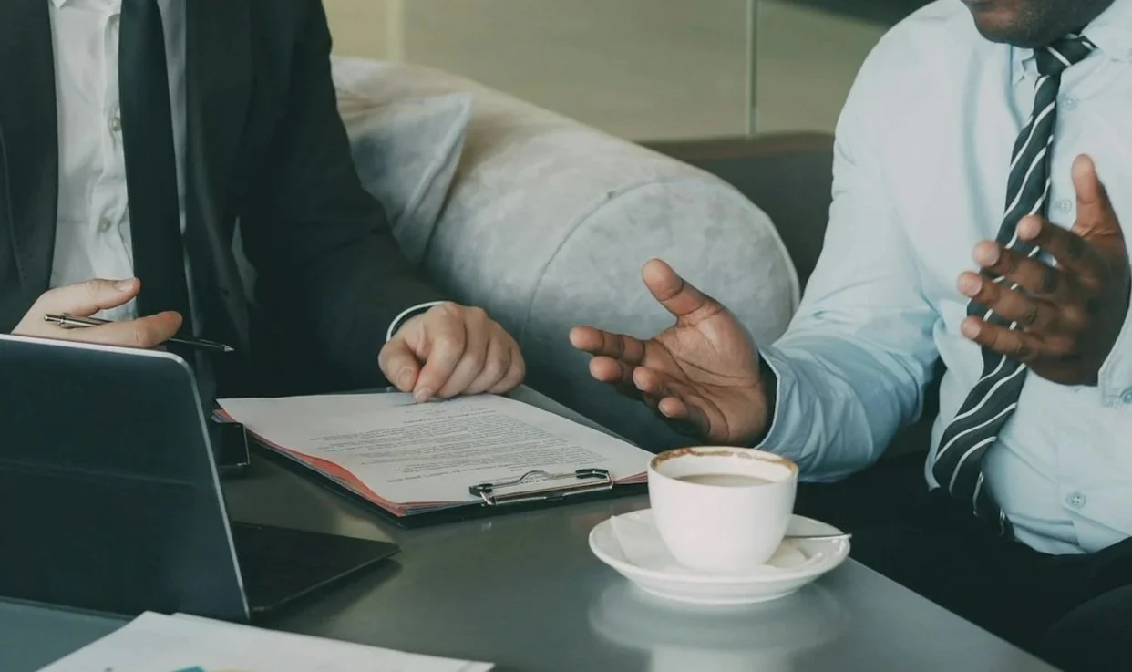 Two men engaged in a business discussion at a table, with a coffee cup and documents in front of them. They appear to be in discussion with each other.