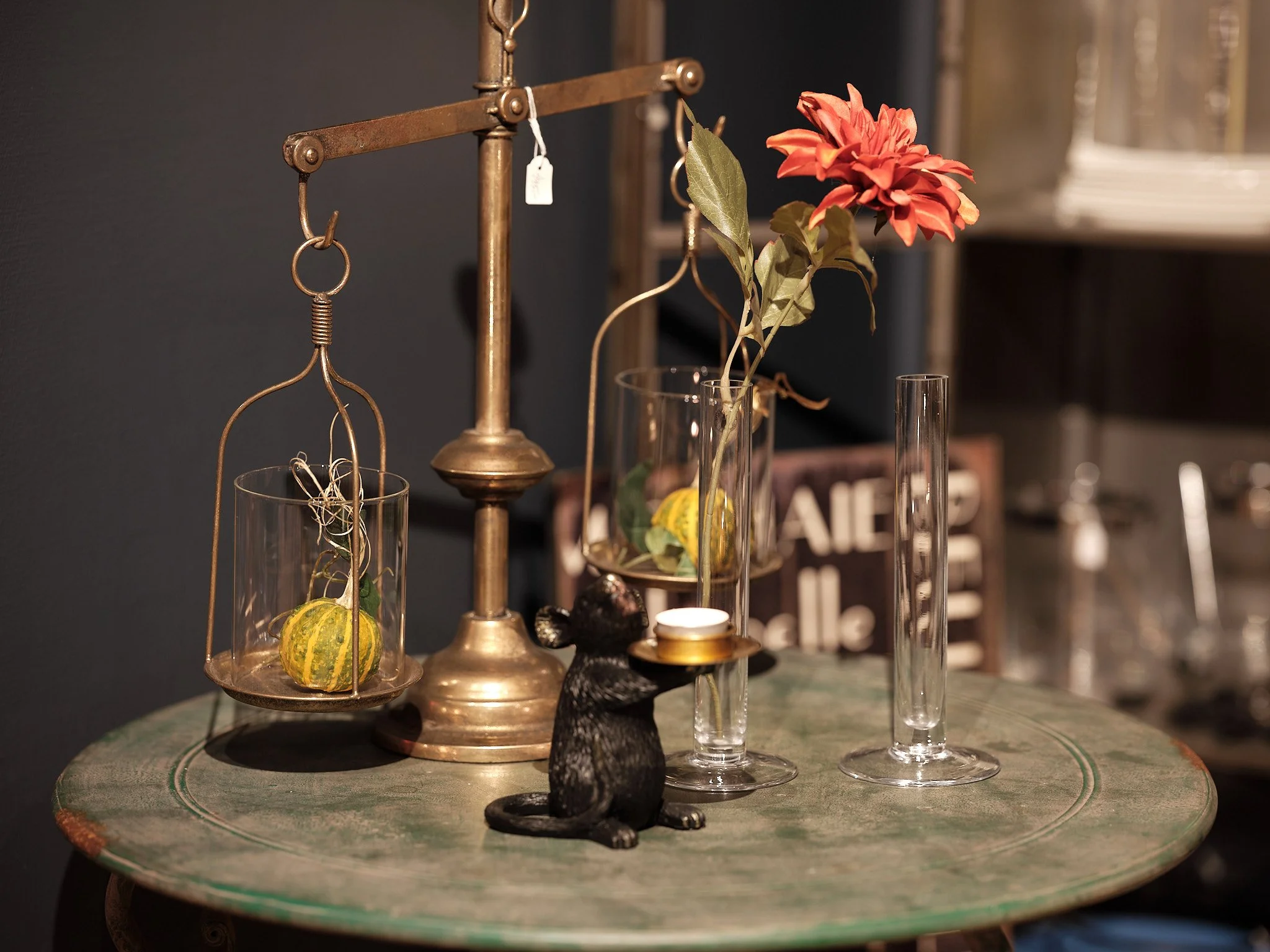 Dekorativ scene med ett grönt bord, ett guldfärgat balanseringsställning med en växt, två tomma glas, en svart kattfigur och ett blomsterarrangemang med en röd blomma