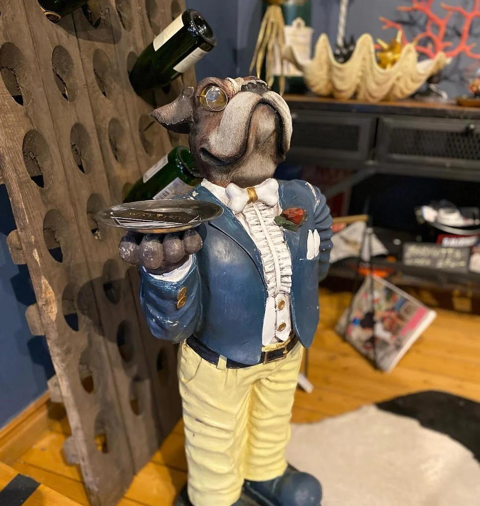 Skulptur av en hund i kostym som håller en bricka med en champagneflaska och glas, klädd i en blå jacket och gula byxor.