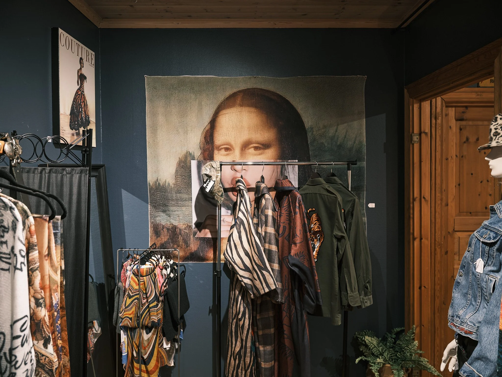 Inredningsdetaljer av en klädbutik med kläder på klädhängare, en väggtextil med ett porträtt av Mona Lisa i bakgrunden och en mannequin i denimjacka till höger.