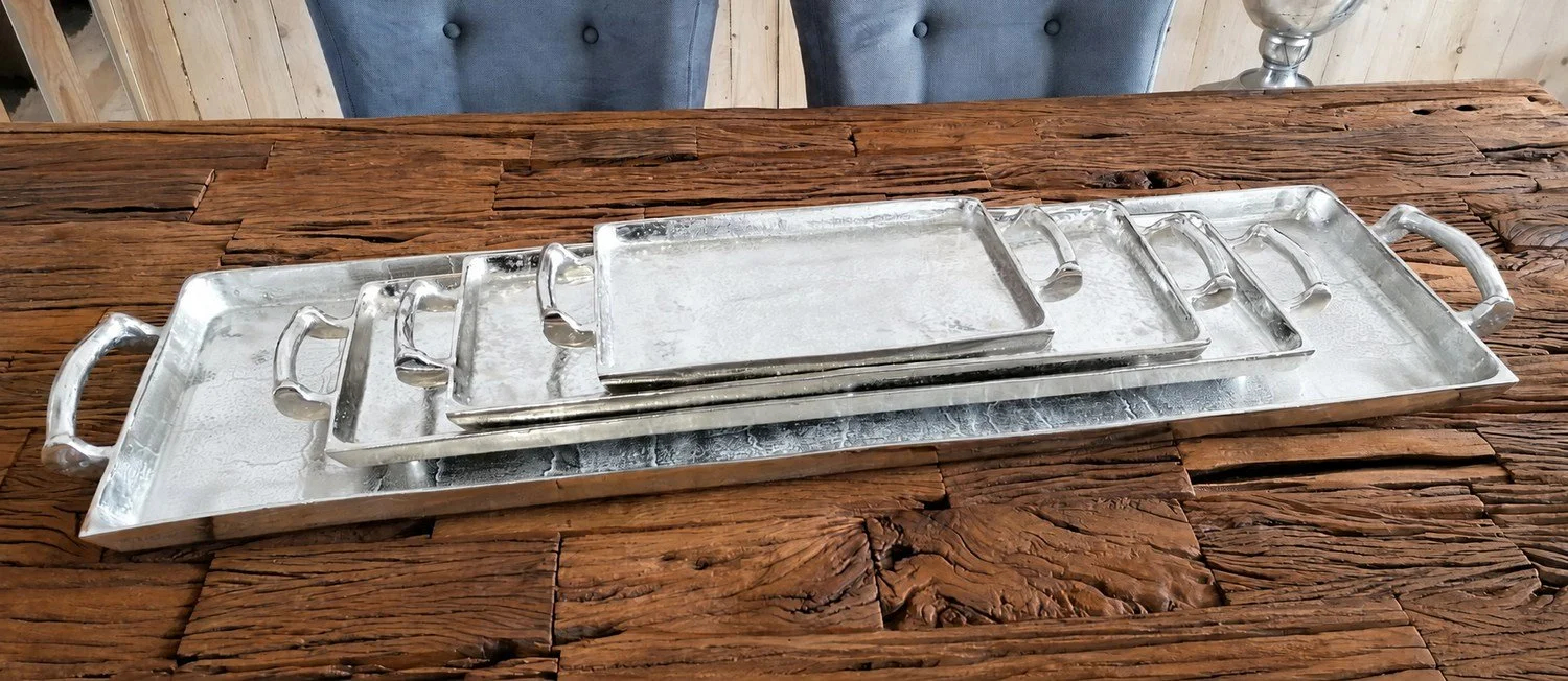Silverfärgade aluminiumbrickor med handtag i flera storlekar på ett träbord. I webbutiken säljs storlek Medium.