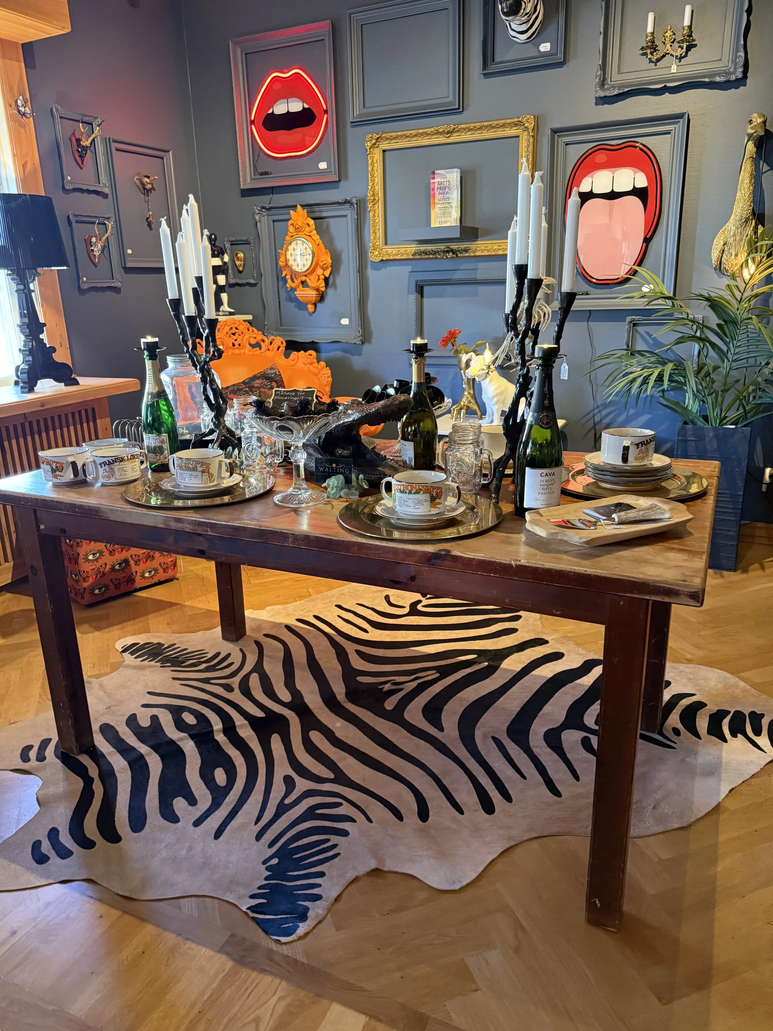 Ett festbord med sex champagneglas, tallrikar, muggar och ljusstakar med vita ljus. Bordsdekorationer inkluderar en gul fågel, en tom glasburk, och ett svart ankarmotiv. Under bordet ligger ett zebra-mönstrat matt- eller skinnmatta. Väggarna är dekorerade med konst, inklusive ansikten med öppna munnar, en gul klocka, och en fågelhuvudskulptur, samt några växter.