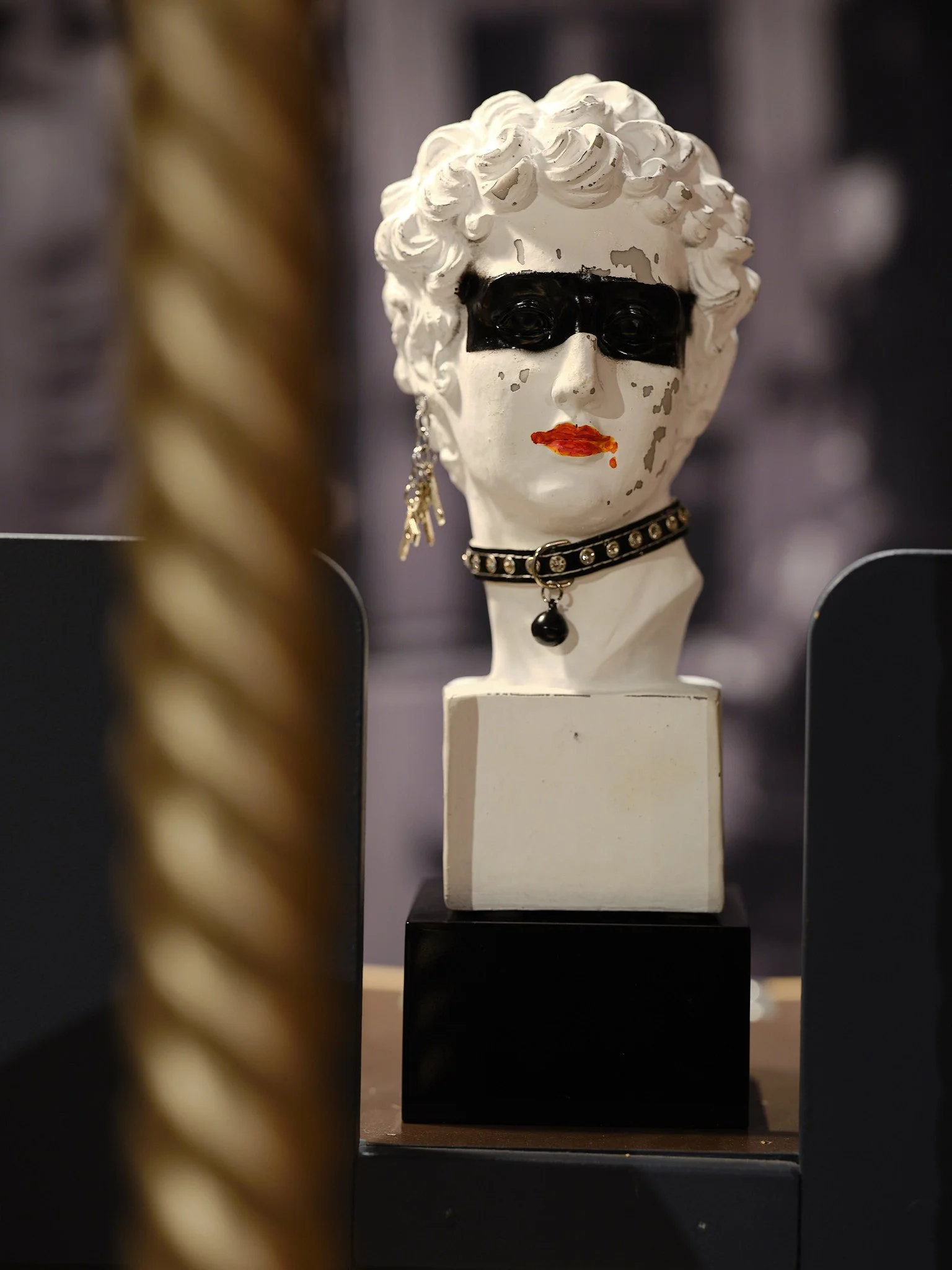 Skulptur av ett konstgjort huvud med svart mask, rött läppstift, en ögonsmycke och ett halsband, placerad på en pedestal.