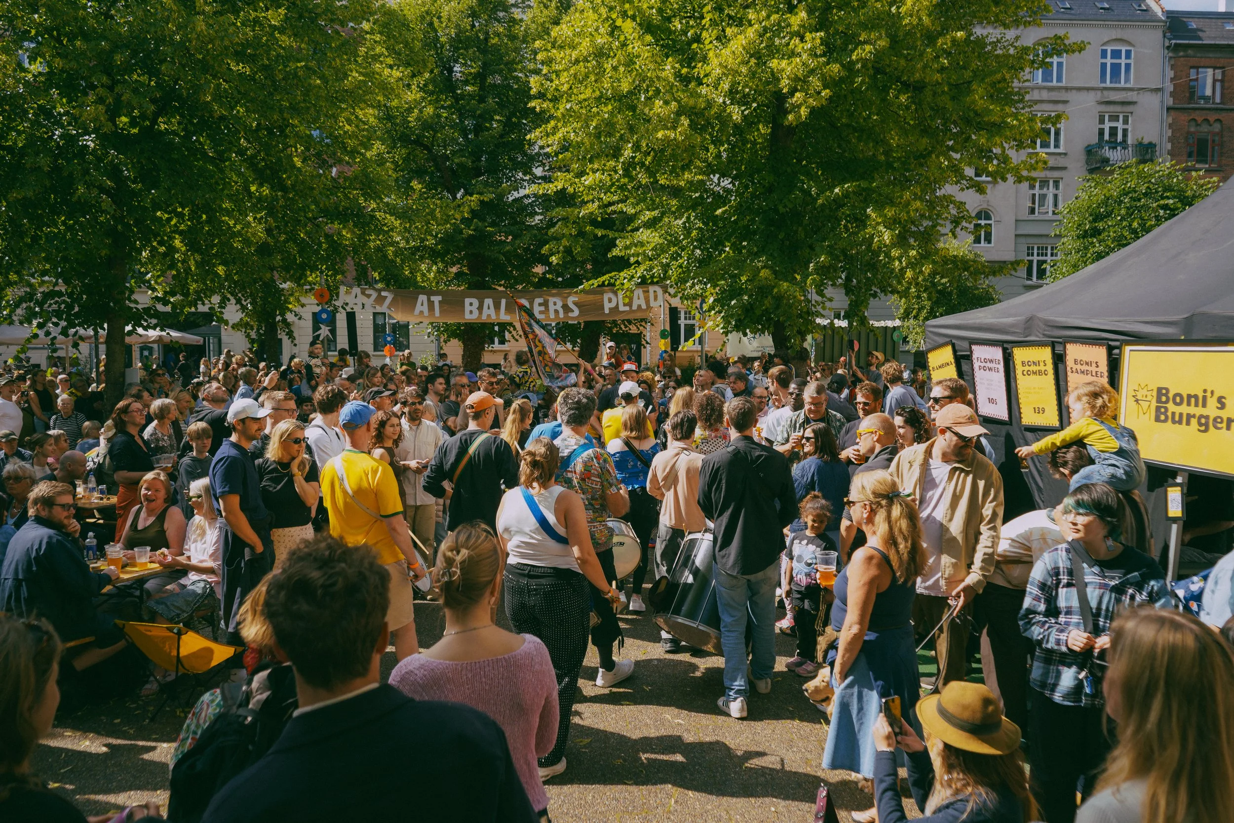 Large crowd gathered outdoors at a festival with green trees and historic buildings in the background. A banner reads 'Jazz at Balzers Platz,' and people are chatting, eating, and enjoying the event.