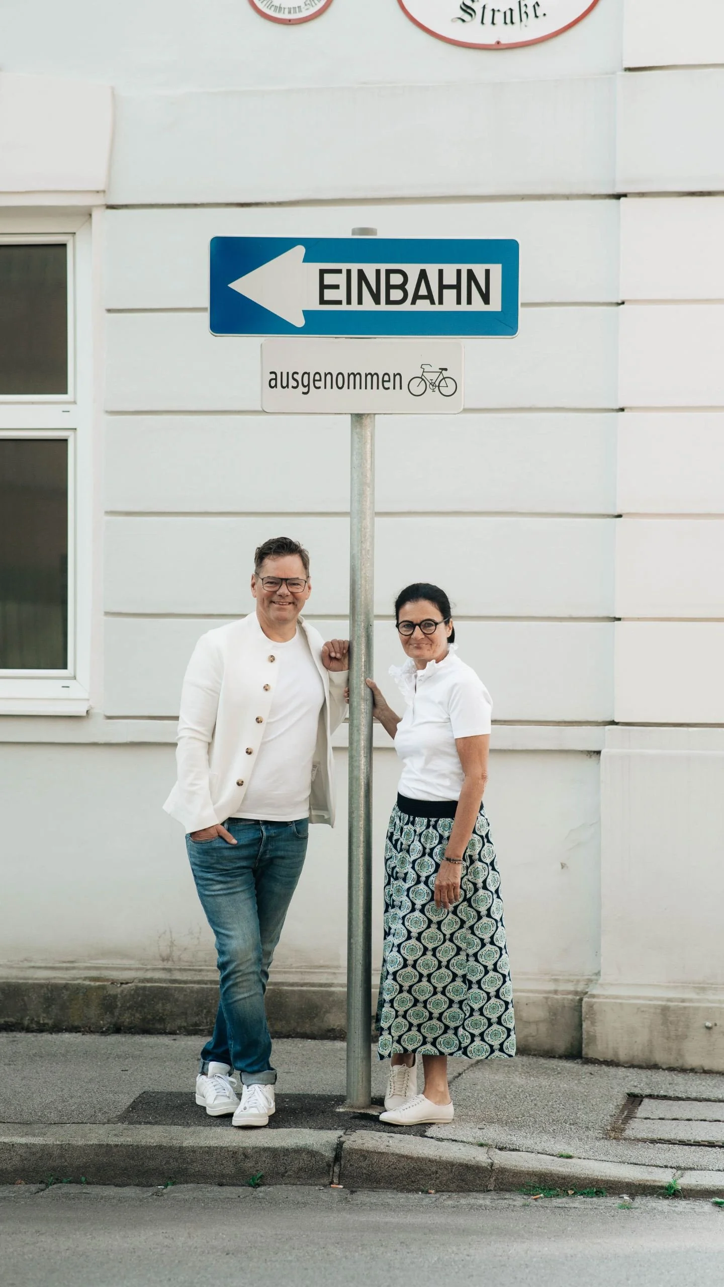 Ein Mann und eine Frau stehen nebeneinander vor einem Straßenschild, das nach links zeigt und mit 'EINBAHN' beschriftet ist. Das Schild hat eine zusätzliche Markierung, die 'ausgenommen' und ein Fahrradsymbol zeigt.
