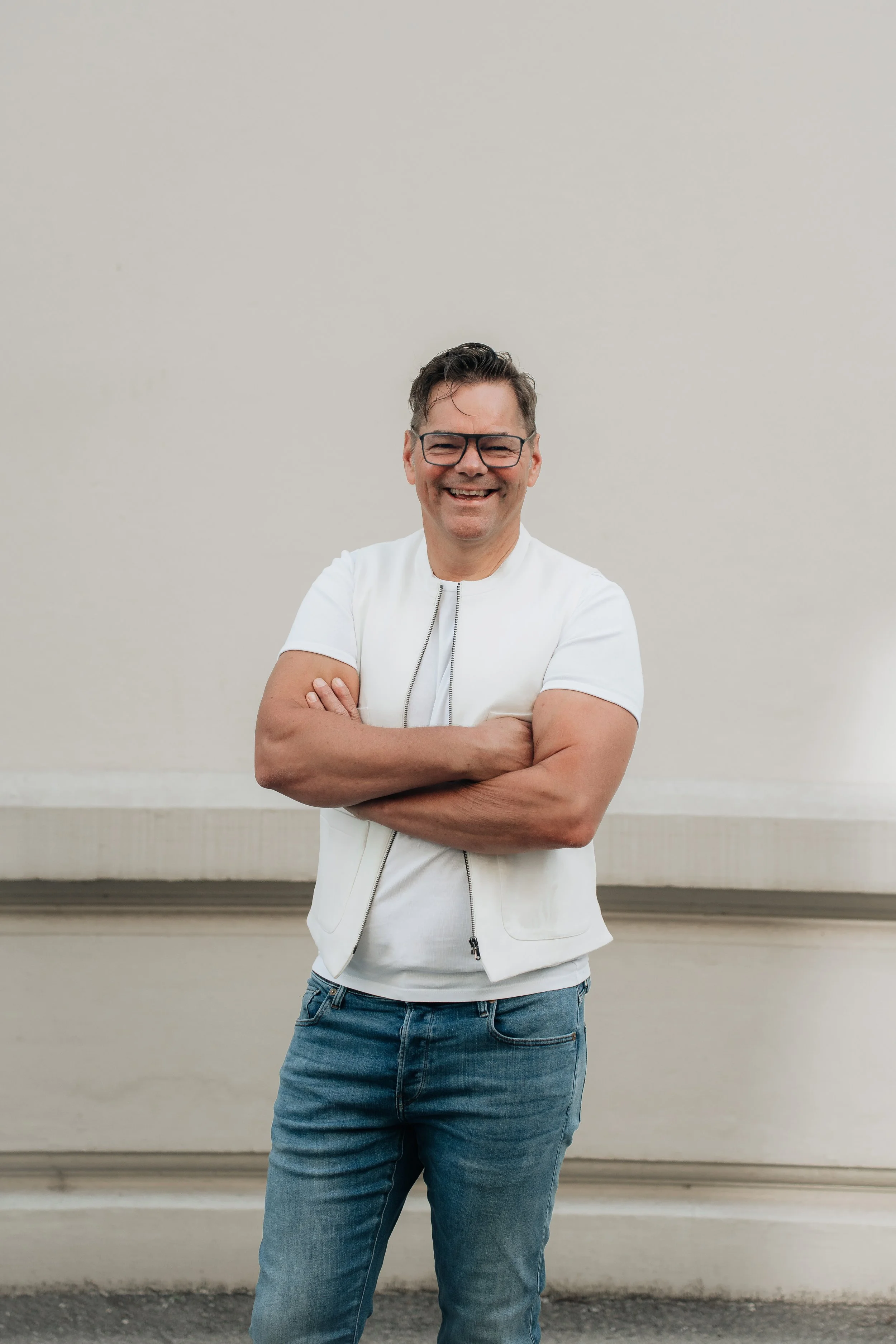 Ein lachender Mann mit Brille, Kurzhaarschnitt, weißen T-Shirt und blauen Jeans steht vor einer weißen Wand.