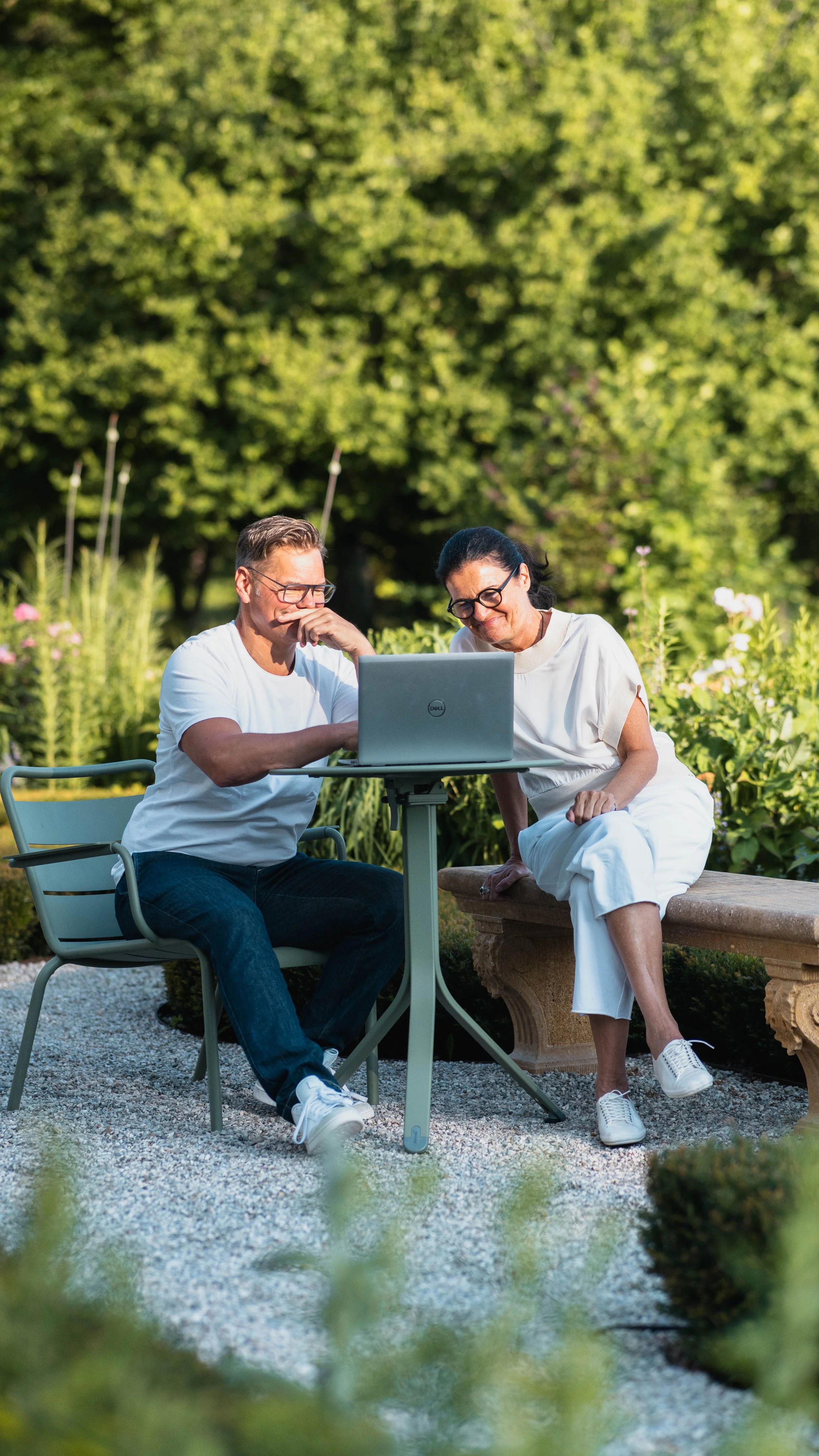 Zwei Menschen sitzen im Garten an einem Tisch, schauen auf einen Laptop, umgeben von grüner Natur und Blumen.