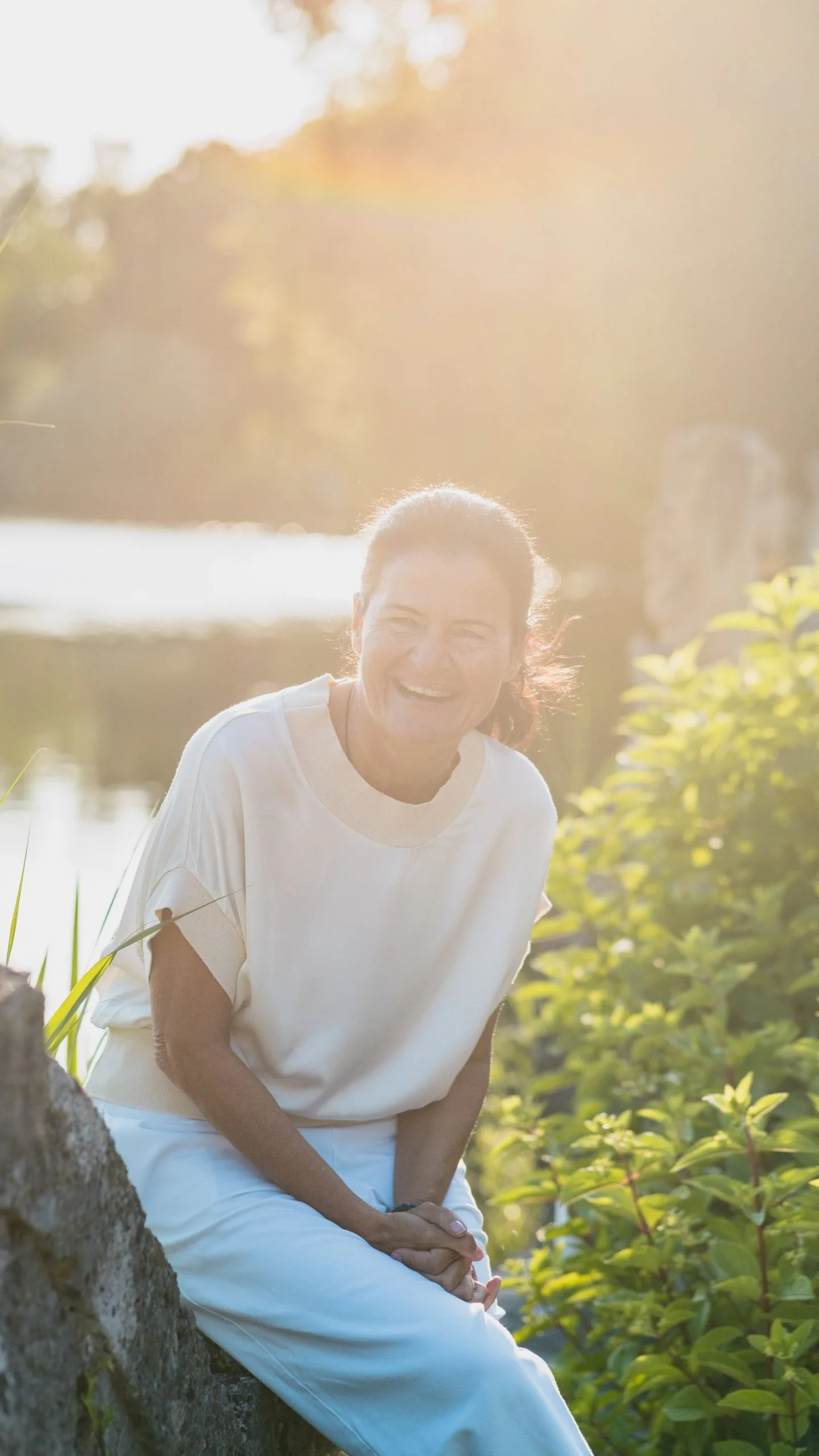 Eine lachende Frau sitzt im Freien bei Sonnenuntergang, umgeben von Natur, an einem Baum. Sie trägt ein weißes T-Shirt und Hosen, die Sonnenstrahlen beleuchten die Szene.