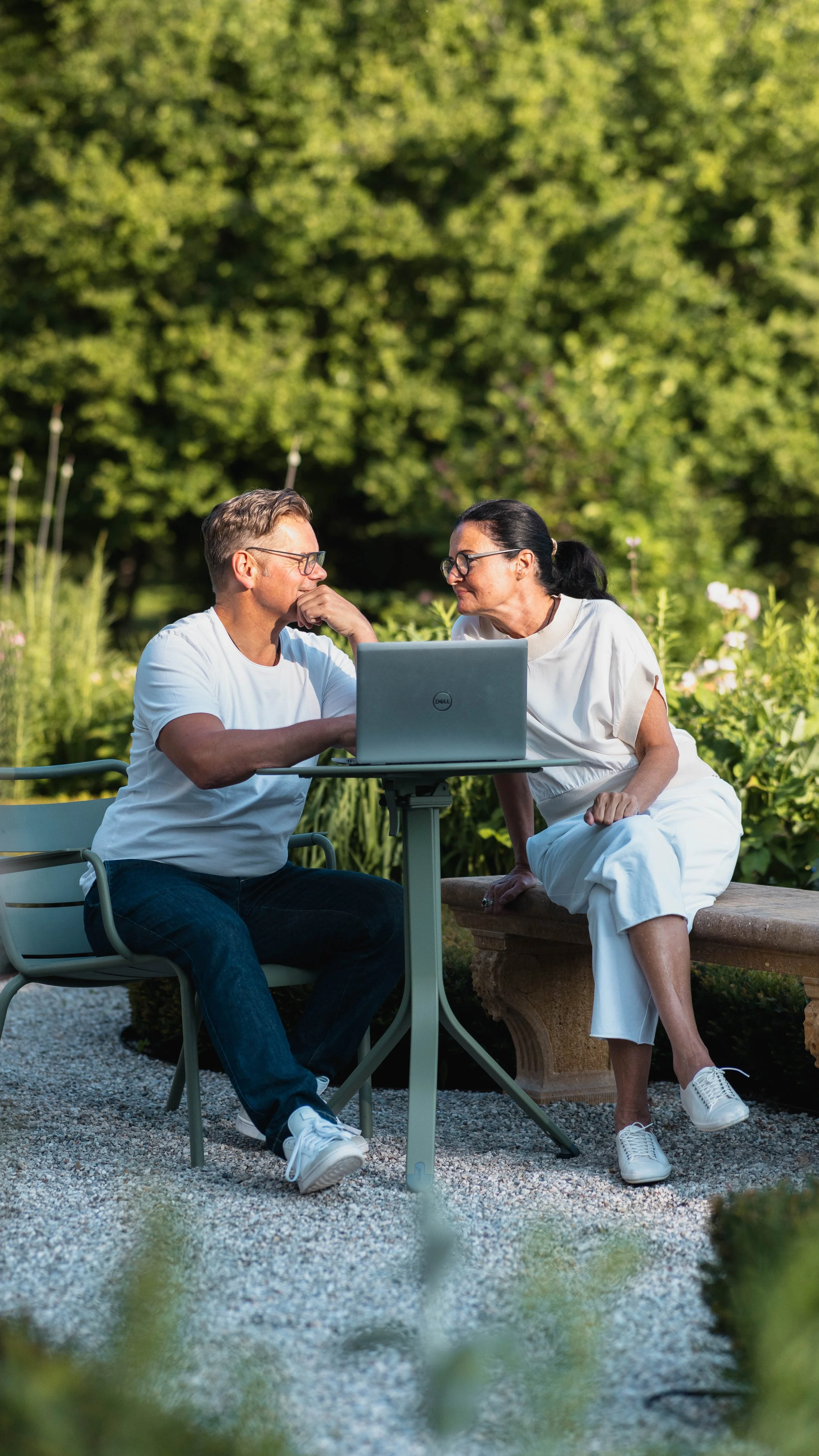 Zwei Menschen sitzen im Freien bei einem Tisch mit einem Laptop, umgeben von grüner Natur, und führen ein Gespräch.