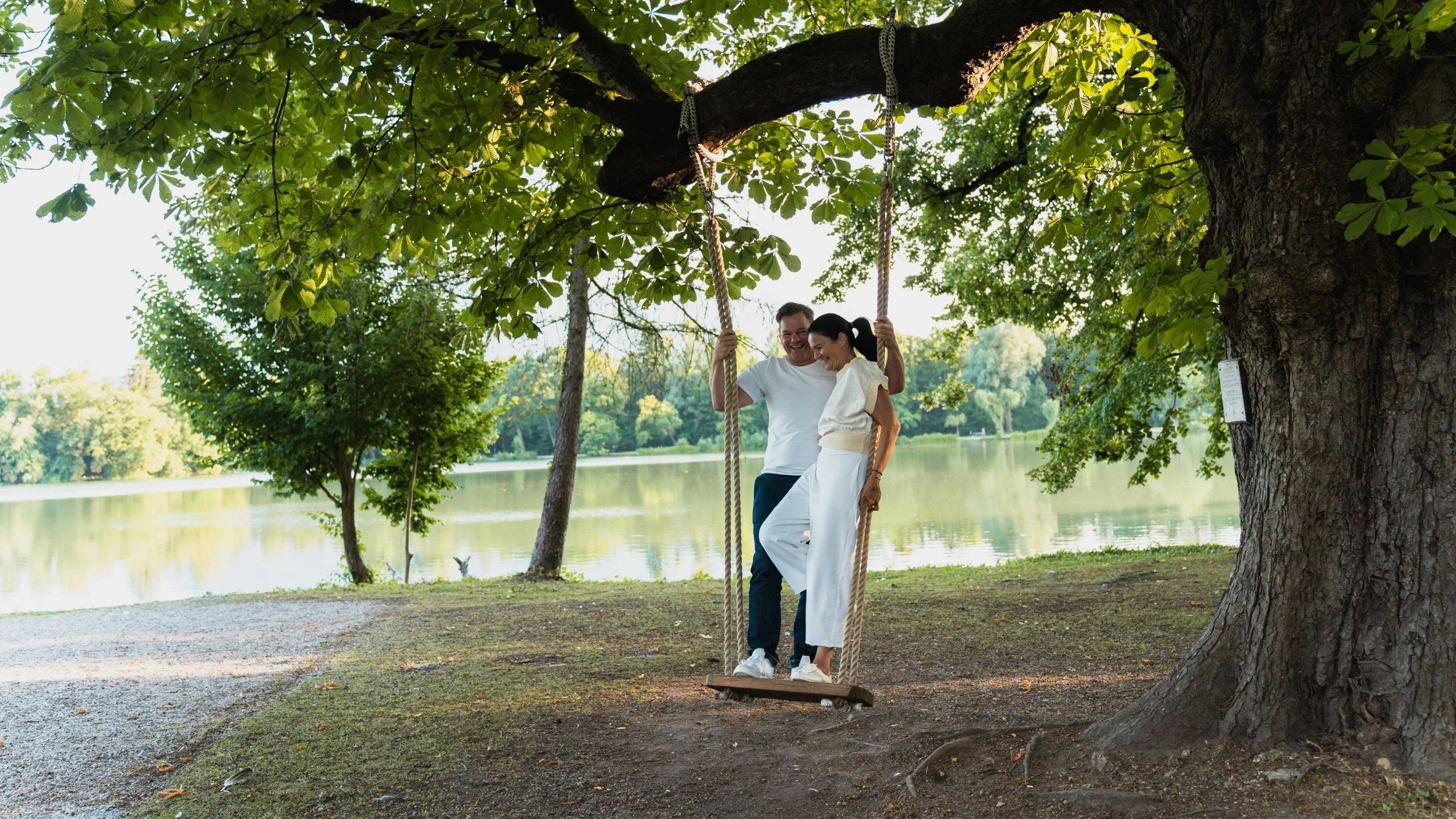 Zwei Menschen sitzen auf einer Schaukel unter einem großen Baum am Fluss, lachen und genießen den sonnigen Tag im Park.