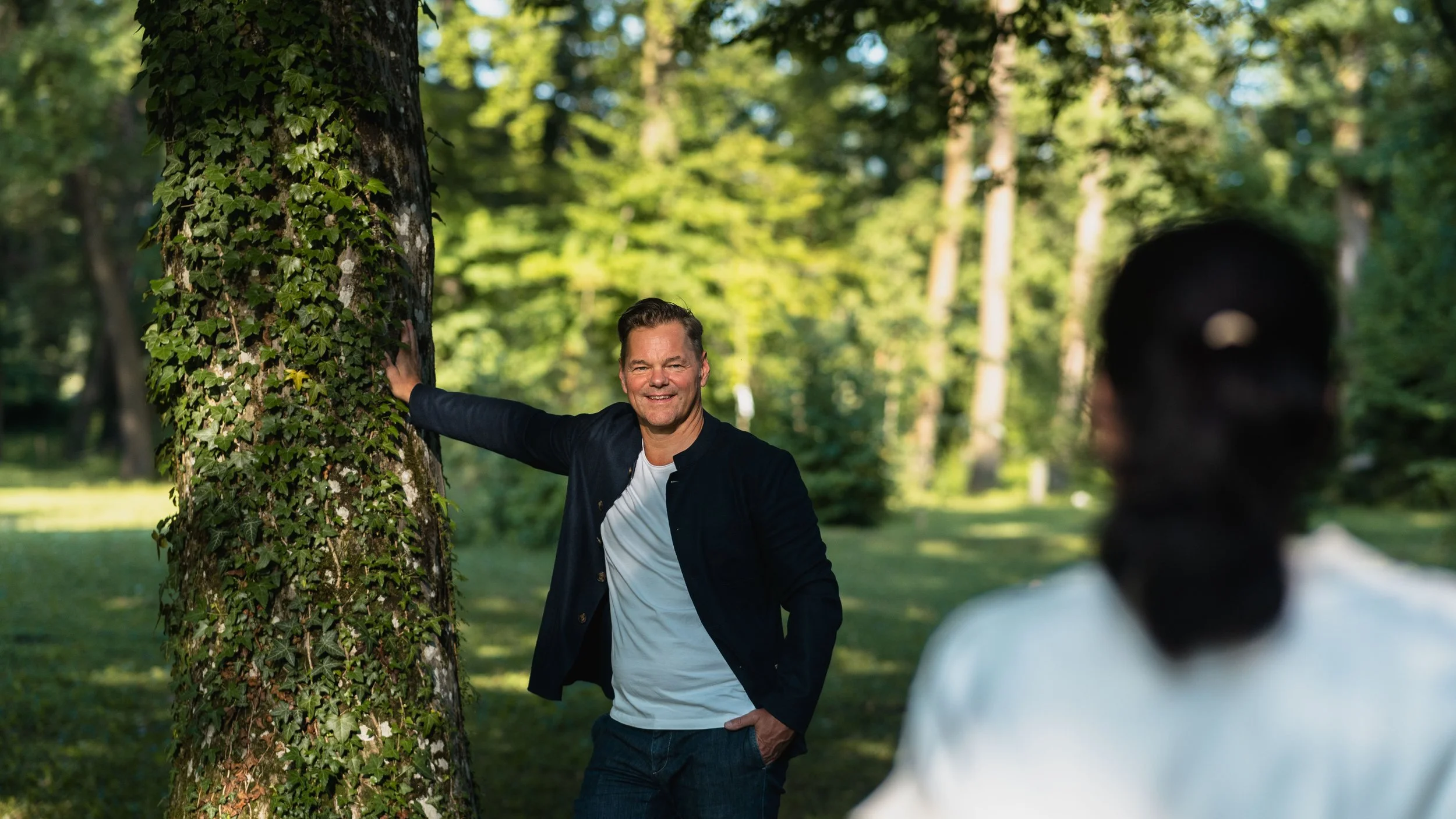Zwei Menschen unterhalten sich im Park, einer lehnt an einem Baum, während die andere Person Blickkontakt hat.
